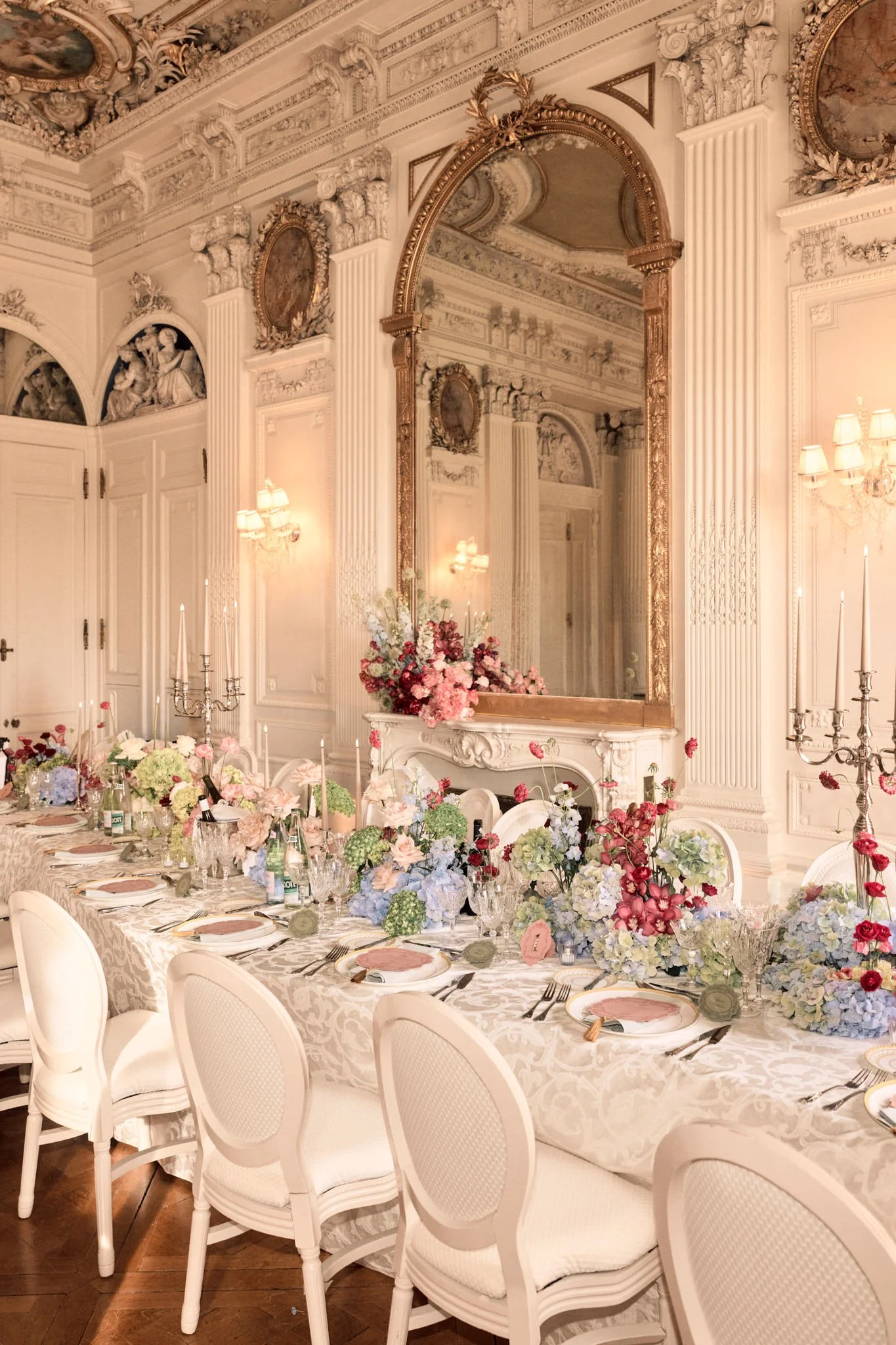 Salle de réception élégante avec table de dîner décorée de fleurs pastel, vaisselle raffinée, bougies et chandeliers, murs ornés de moulures et un grand miroir doré, ambiance chaleureuse et sophistiquée.