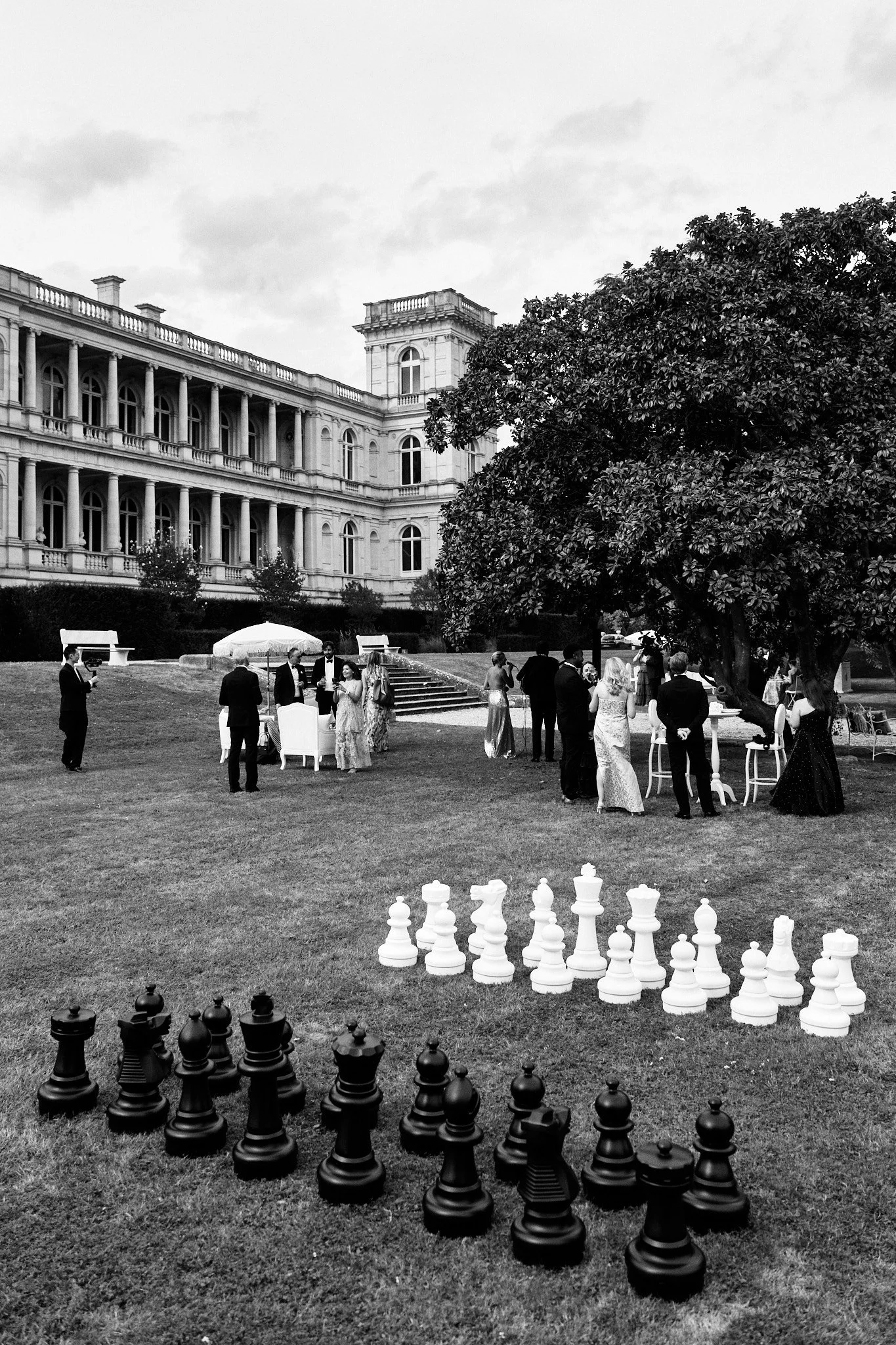 Un groupe de personnes habillées élégamment, jouant aux échecs géants dans un jardin devant un grand bâtiment historique, avec un arbre imposant à droite.