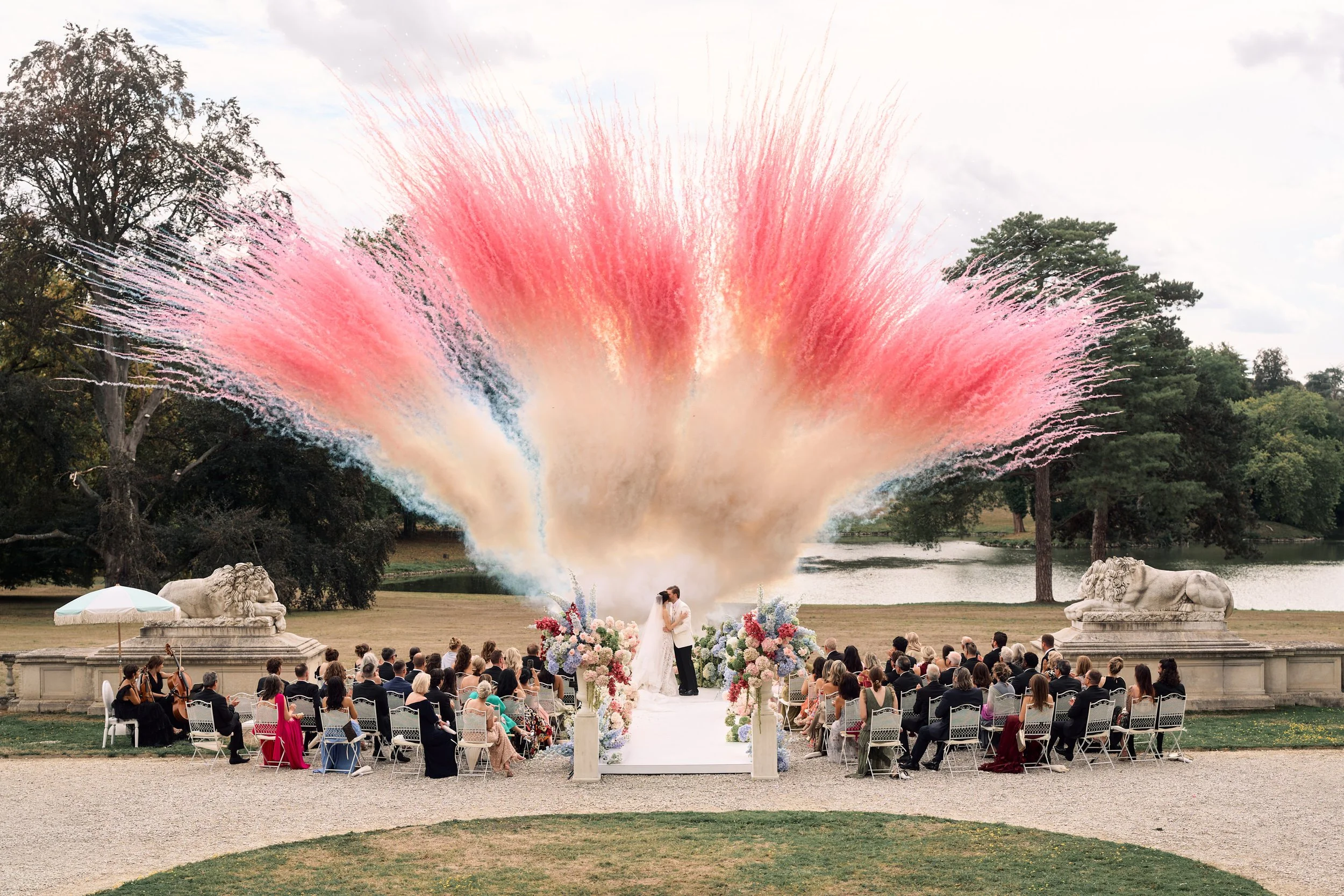 Un mariage en plein air avec un feu d'artifice rose au-dessus, un couple de mariés s'embrassant devant une foule d'invités, avec un décor floral élégant et deux statues de lions, près d'un lac et entouré d'arbres.