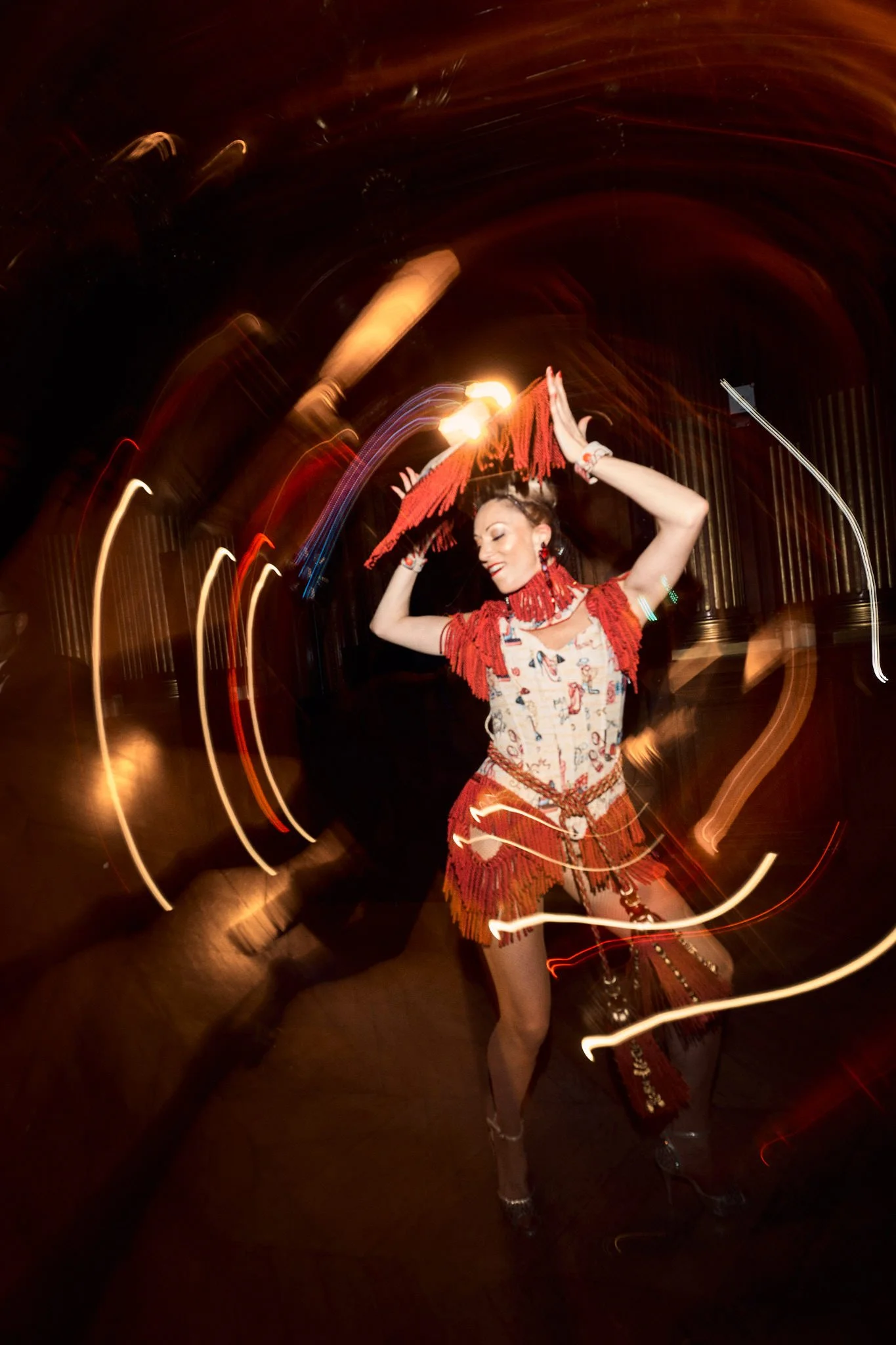 Une femme danse lors d'une performance, portant un costume coloré avec des franges rouges et des accessoires en perles. L'image est prise avec un effet de mouvement, créant des trails de lumière.