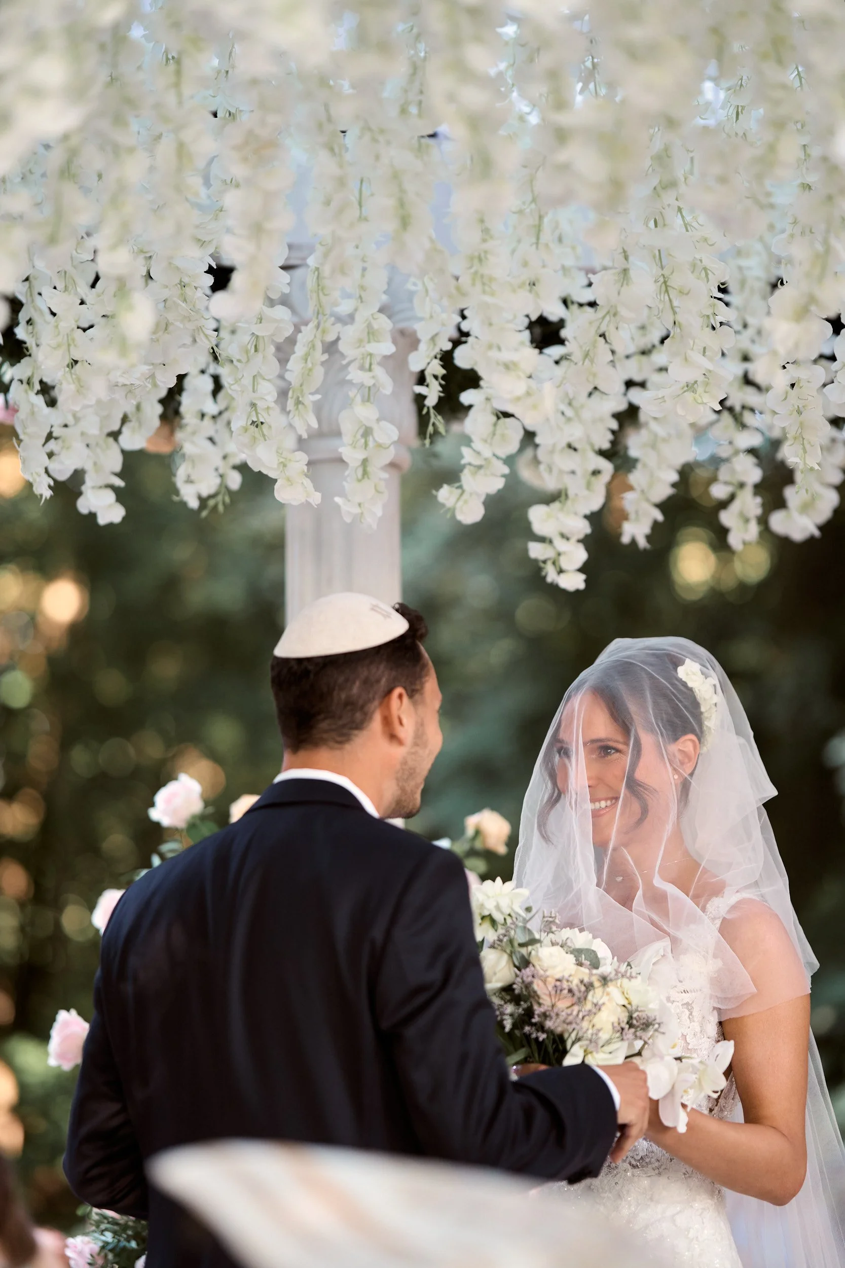 Un couple de mariés échangeant leurs vœux lors de leur mariage en extérieur, sous une décoration florale blanche avec un voile et des fleurs.