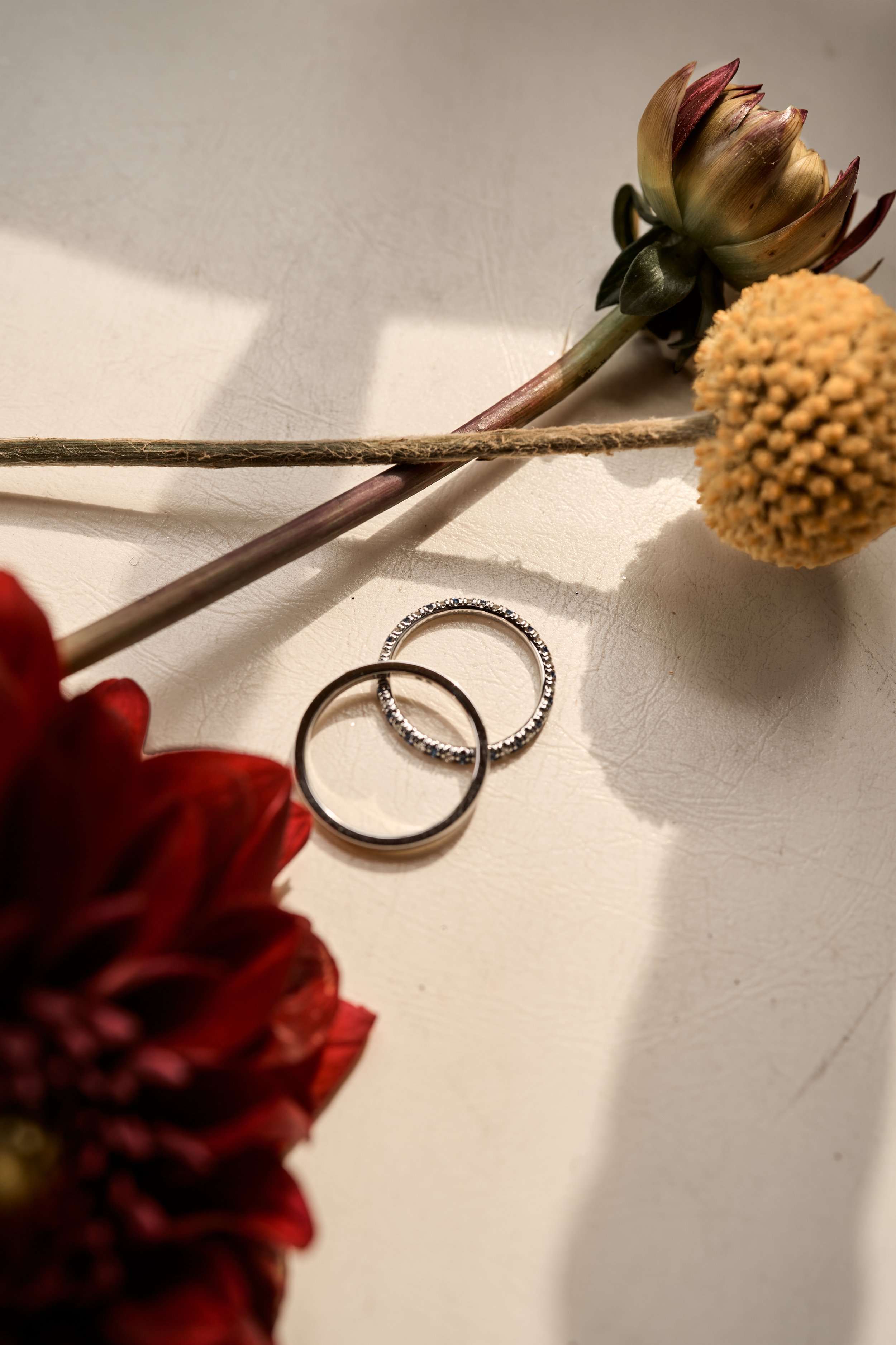 Deux alliances en argent, une lisse et une ornée de petits diamants, posées sur une surface blanche, entourées de fleurs sèches rouges et jaunes.
