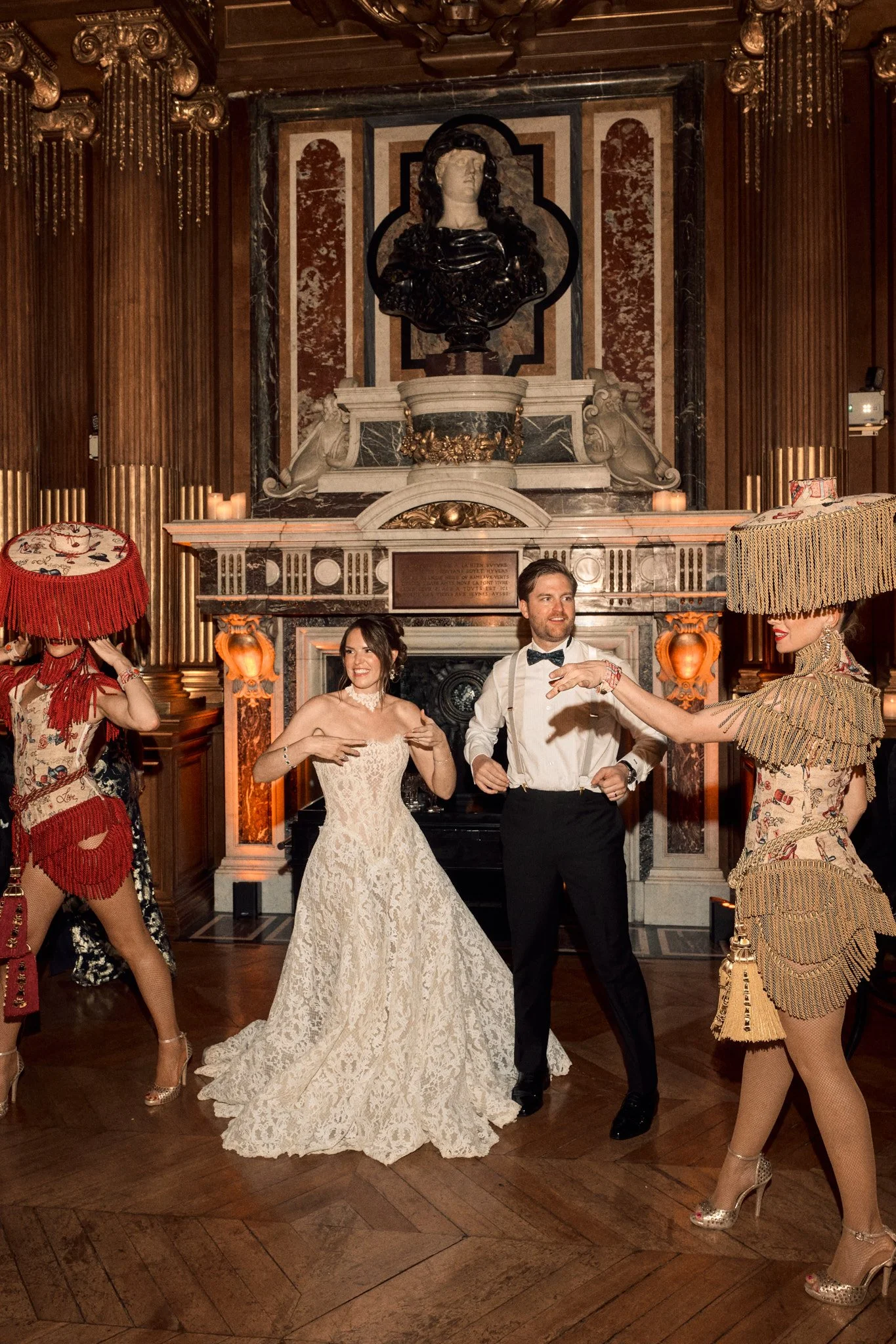 Une groupe de personnes en réception ou soirée élégante, avec une femme en robe de luxe et d'autres femmes portant des chapeaux décoratifs, dans une pièce richement décorée avec un grand portrait en arrière-plan.