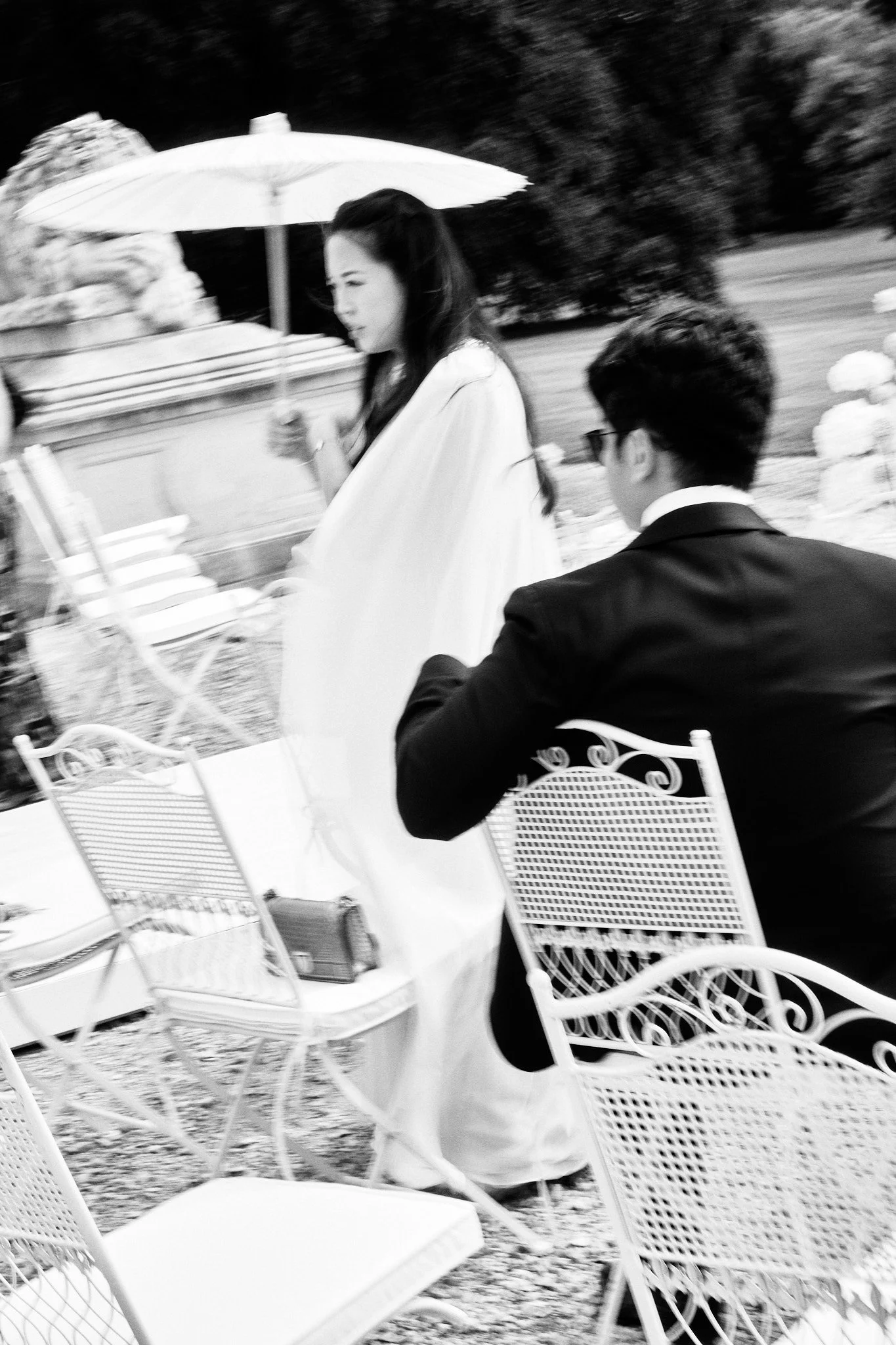 Une femme avec un parapluie blanc lors d'un événement en plein air, assise à une table à l'extérieur, près d'une autre personne avec des cheveux bouclés, en noir et blanc.