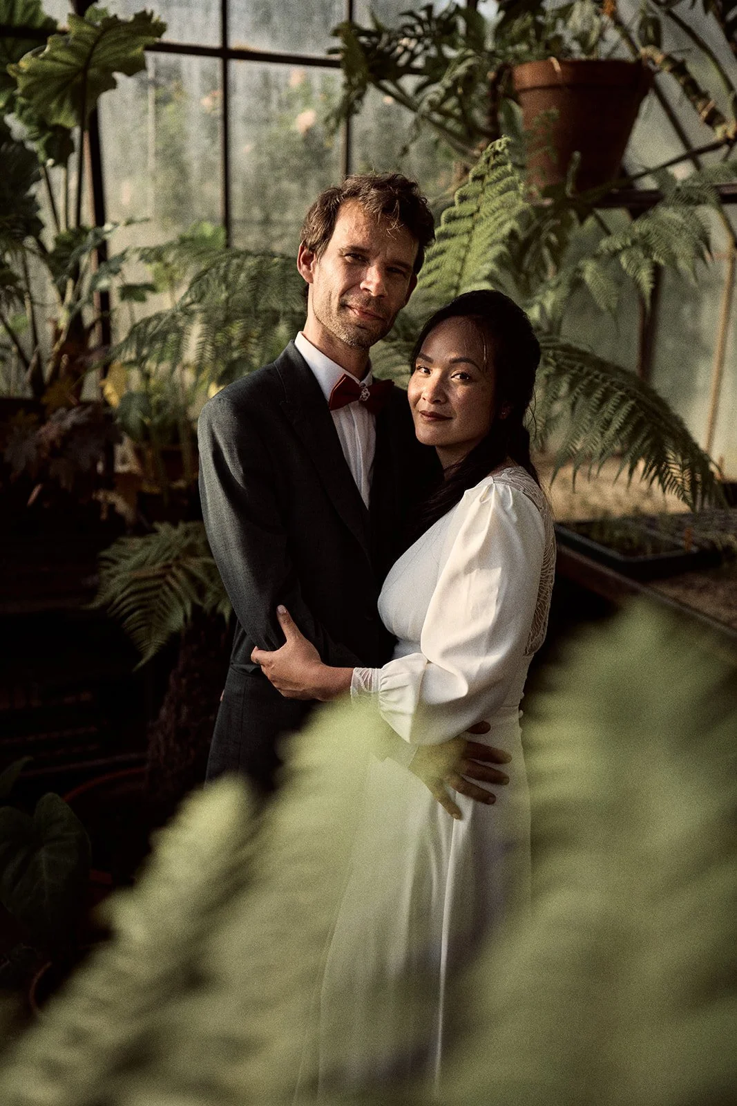 Un couple en costume habillé pour une occasion spéciale, en train de s'embrasser dans une serre remplie de plantes vertes.