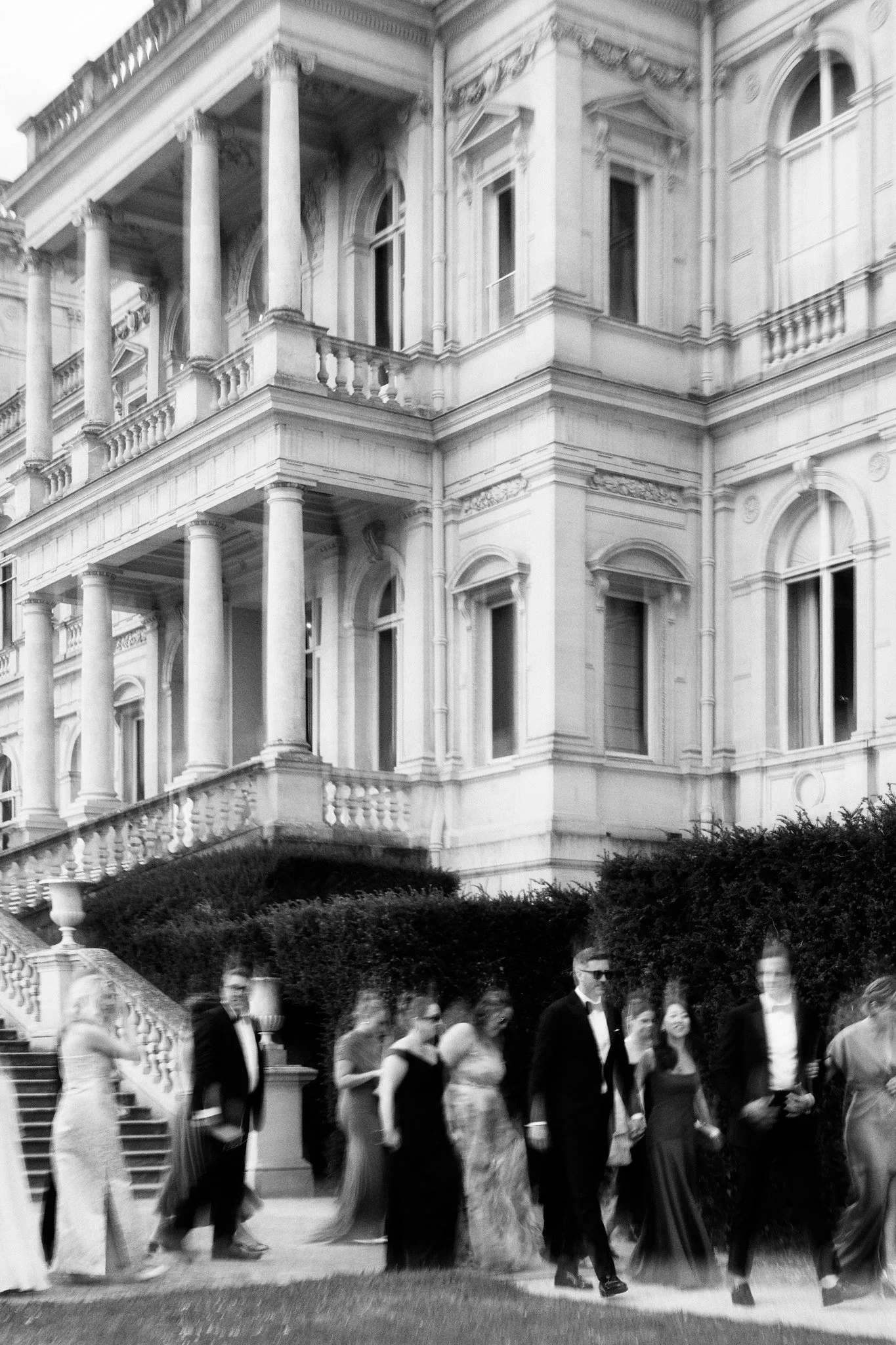 Un groupe de personnes en tenue formelle marchant en procession devant un bâtiment historique avec des colonnes et des balustrades, probablement lors d'un événement officiel ou d'une cérémonie.