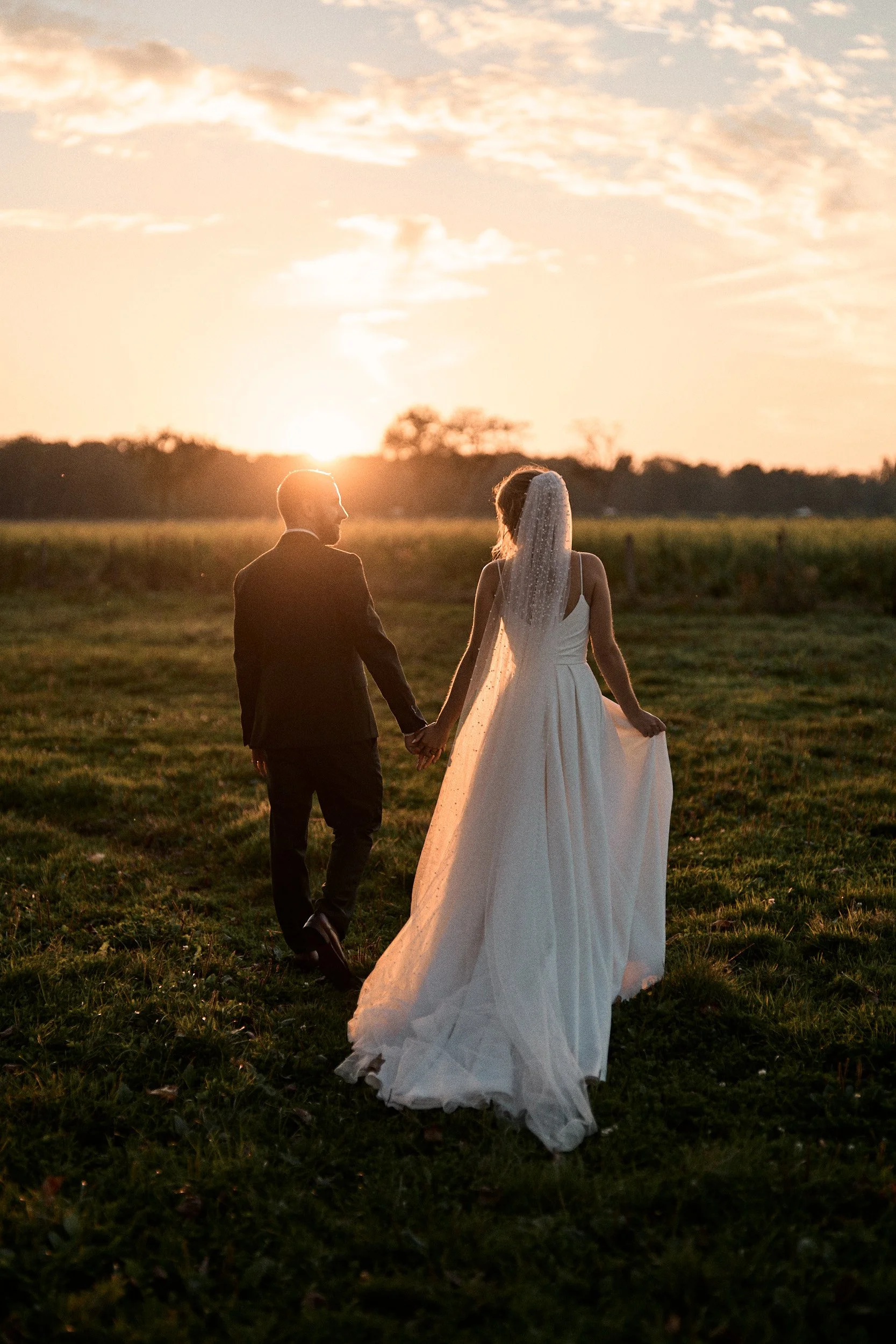 Un couple de mariés marche main dans la main dans un champ au coucher du soleil, la femme porte une robe de mariée blanche avec un voile et l'homme porte un costume noir.