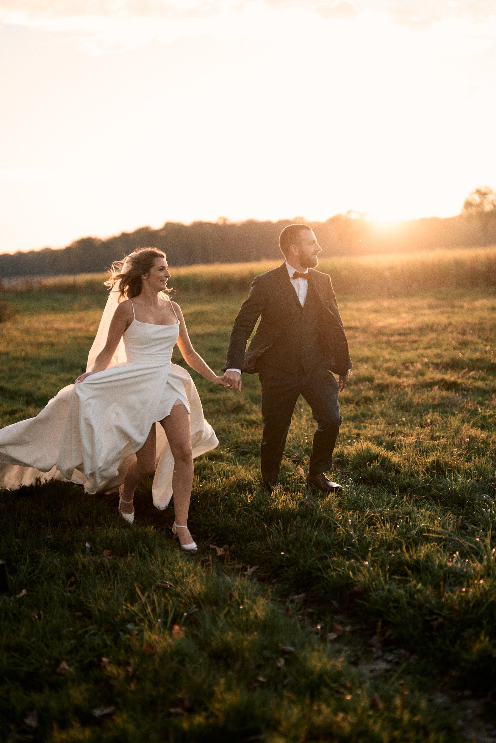 Un couple en tenue de mariage qui marche dans un champ lors du coucher du soleil, se tenant par la main.