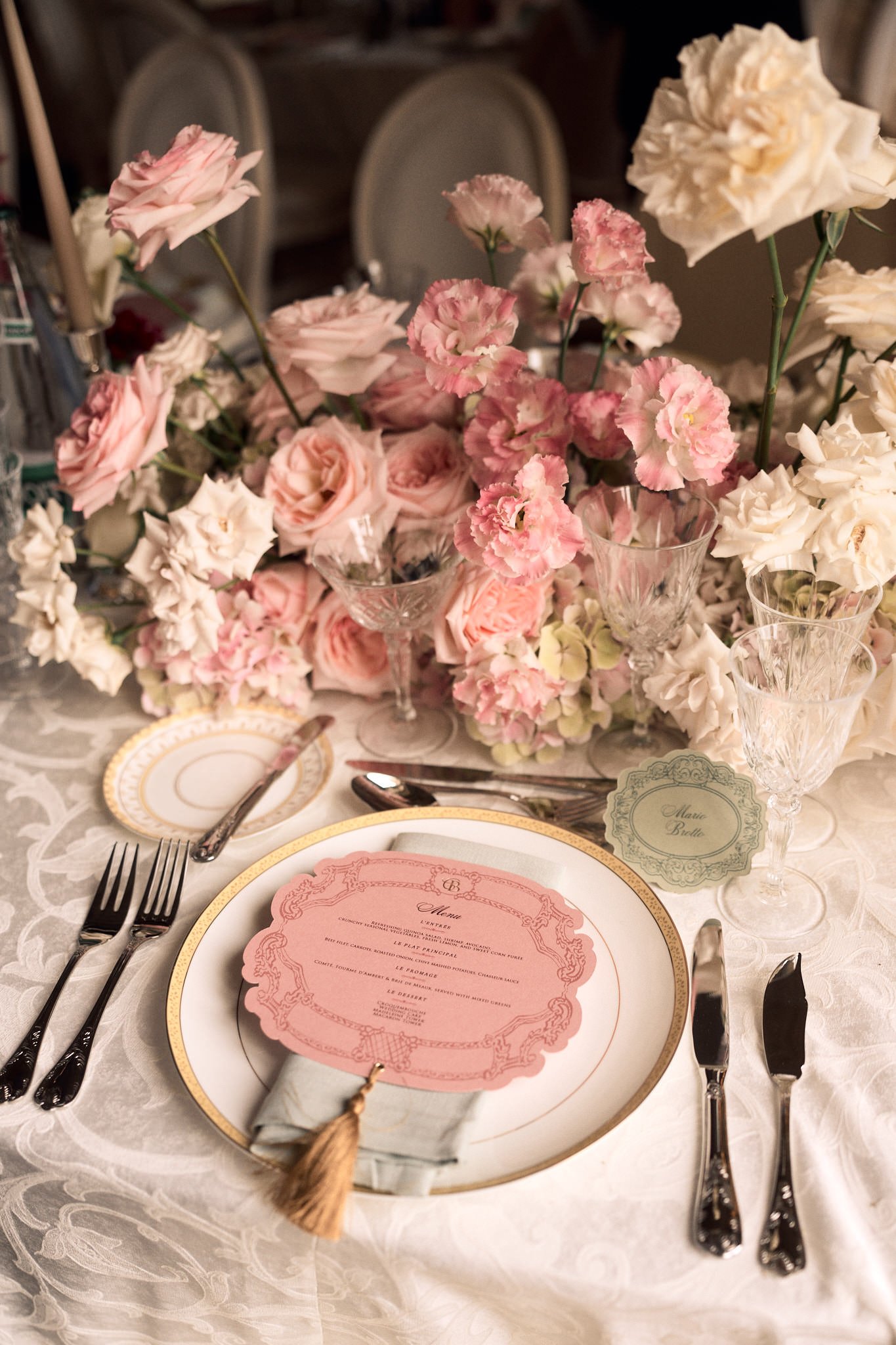 Décor de table romantique avec un arrangement floral de roses roses et blanches, un menu rose en papier avec un attaché-serrure et une pampille en ficelle, assiettes en porcelaine blanche avec un motif doré, verres à vin en cristal, et couverts en mé