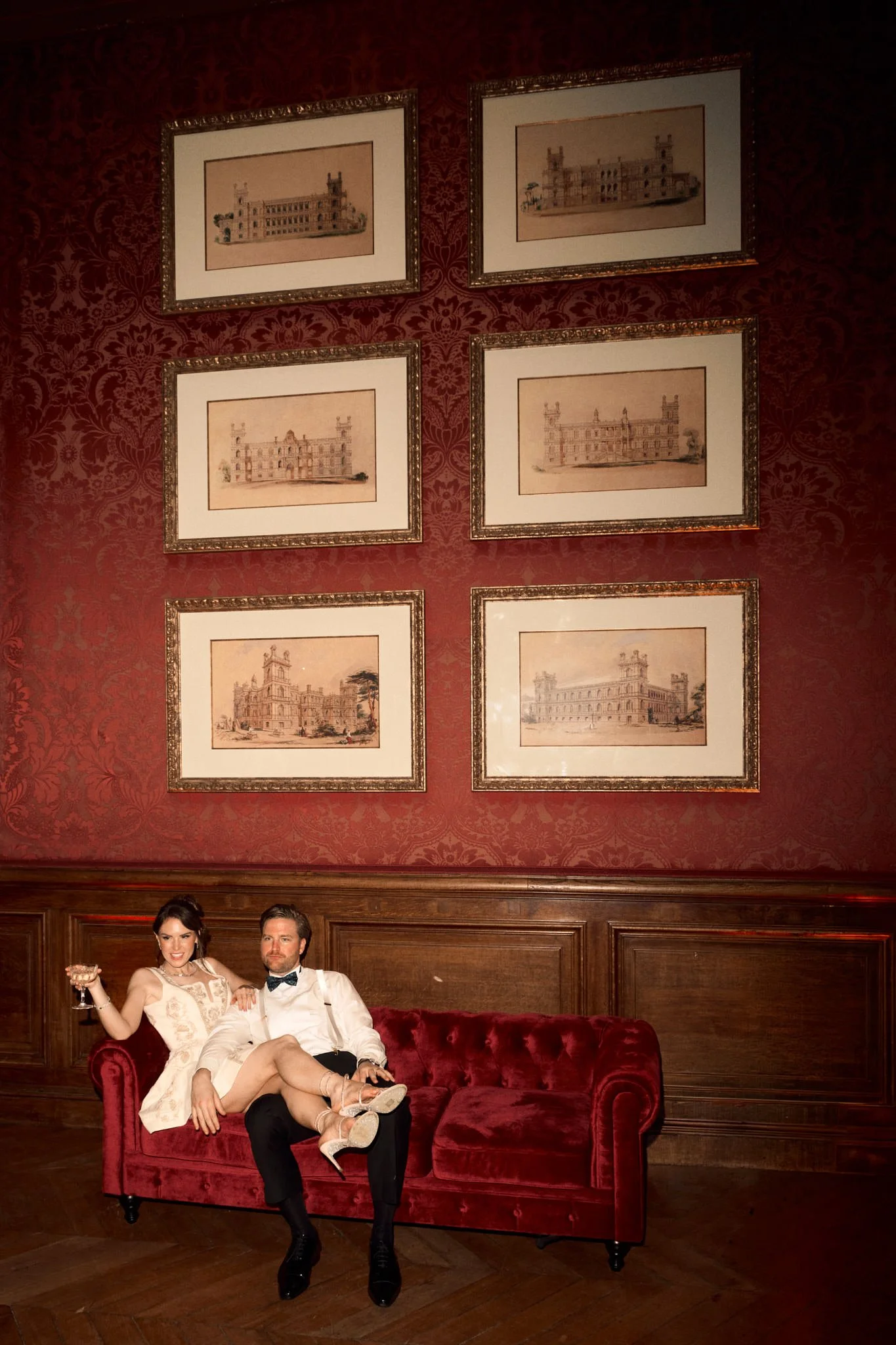 Un couple assis sur un canapé rouge dans une pièce élégante avec des murs ornés de cadres contenant des dessins de château.