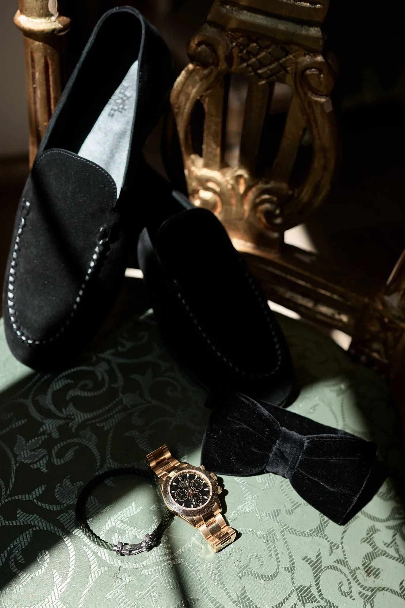 Une montre en or avec un bracelet en métal, une paire de chaussures noires avec un nœud en velours, une montre noire, un bracelet en acier et un autre en cuir, une clé, une veste en velours noir, un oreiller en velours noir, et un fond en tissu vert 