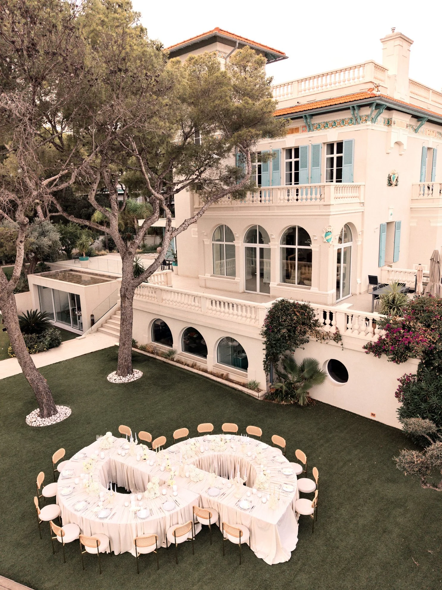 Une maison élégante avec une terrasse, entourée d'arbres et de plantes, avec une table ronde décorée pour un événement en plein air.