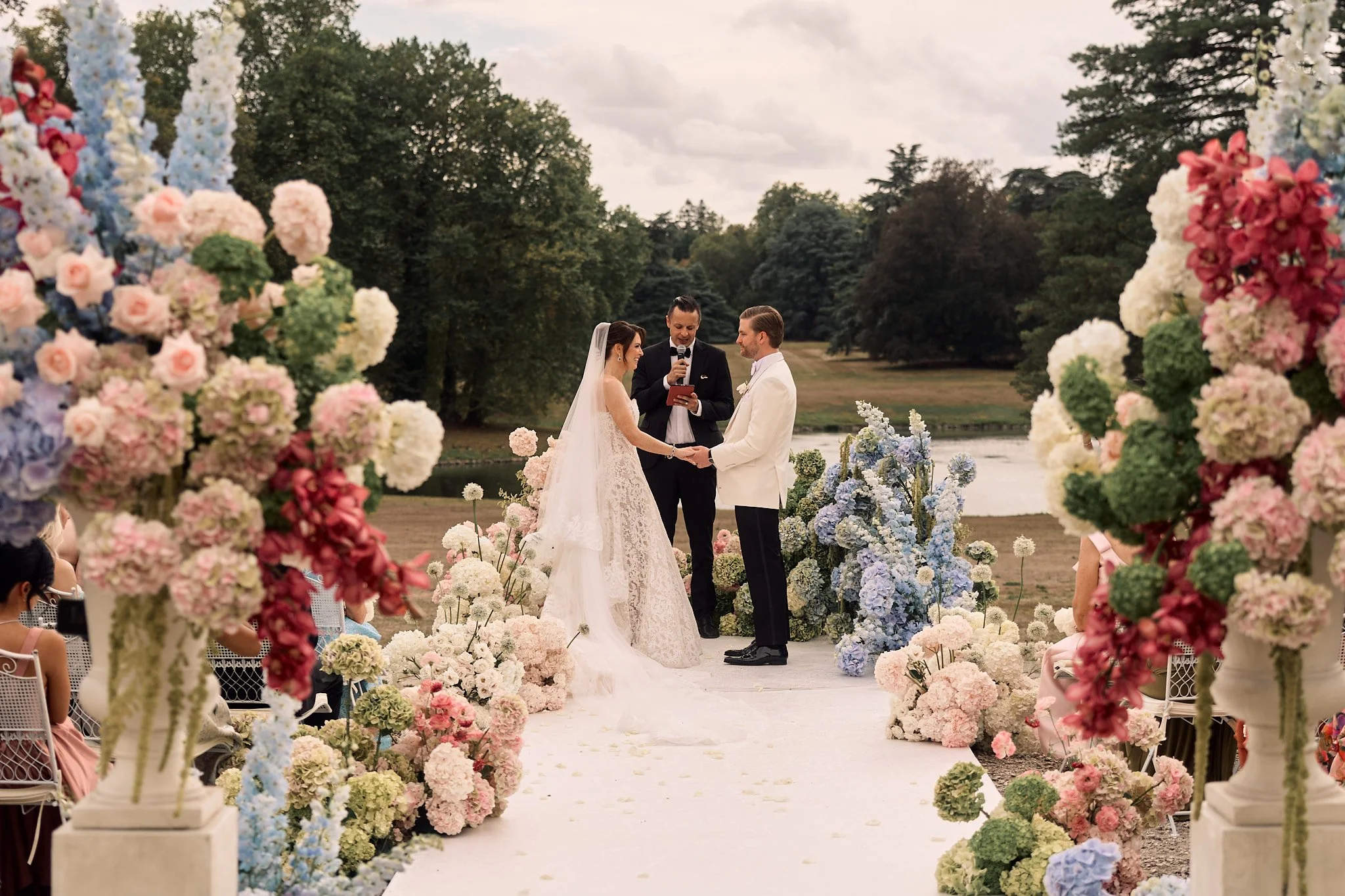 Un couple lors de leur mariage debout devant un officiant, entourés de fleurs colorées avec un décor naturel au bord d'une rivière.