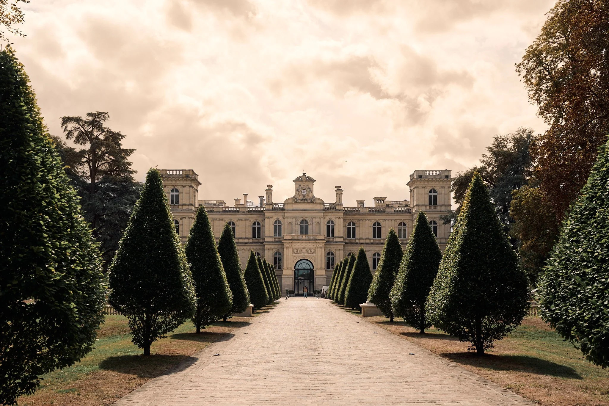 Un palais ancien avec une façade ornée, entouré d'un jardin avec une allée bordée d'arbustes taillés, sous un ciel nuageux.