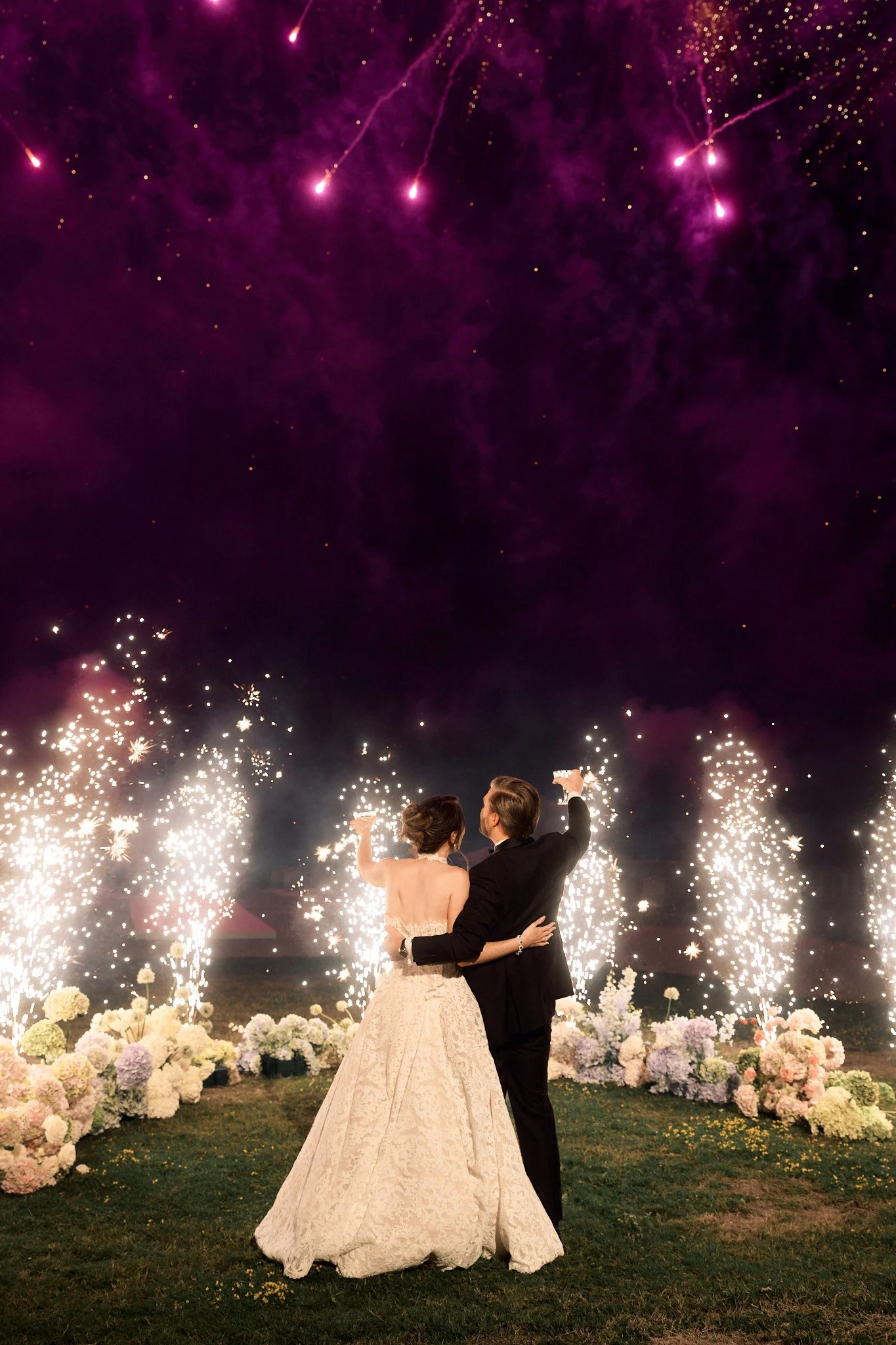 Un couple en robe de mariage et costume, vu de dos, célébrant leur mariage avec des feux d'artifice colorés dans le ciel et entourés de fleurs, dans une scène nocturne romantique.
