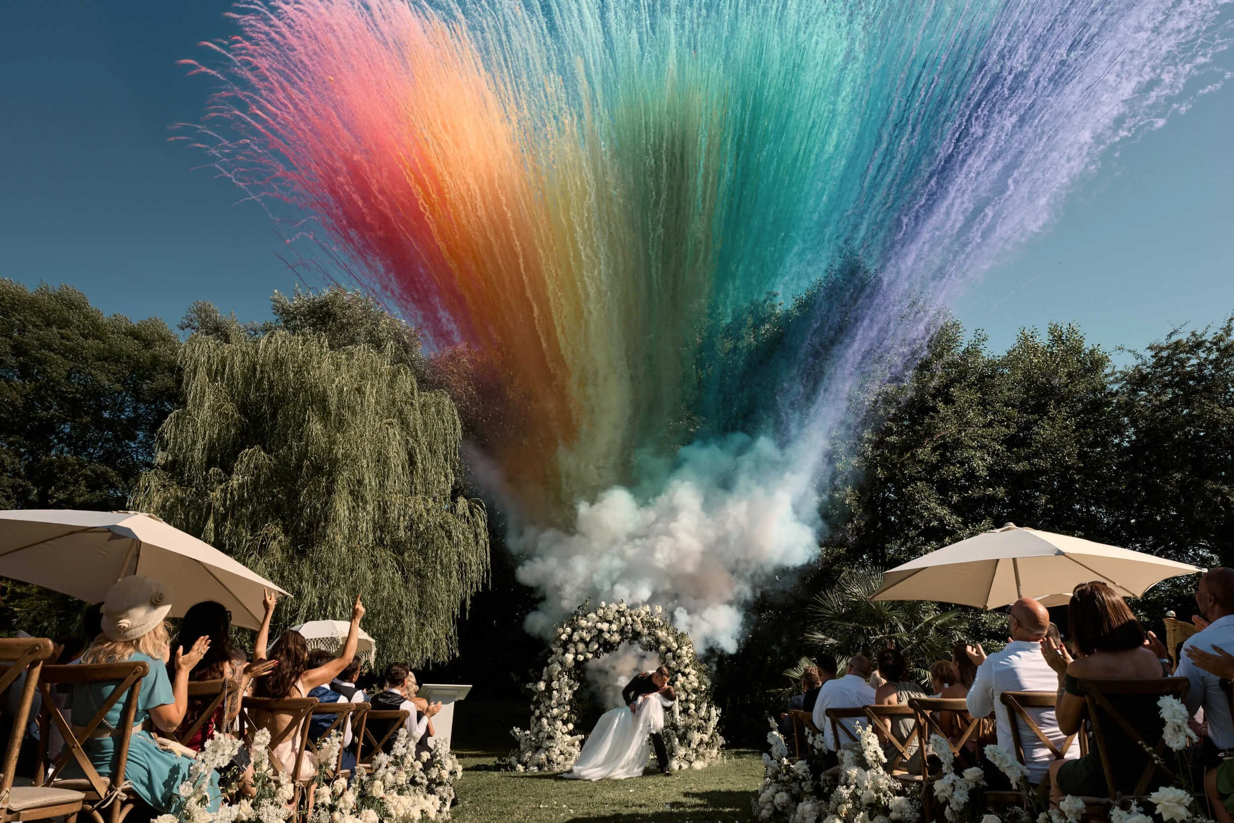 Un grand groupe de personnes assises dans un jardin, regardant un feu d'artifice en forme d'arc-en-ciel dans le ciel bleu. La scène semble être une célébration ou un mariage en plein air, avec des chaises en bois, des parasols et des décorations flor