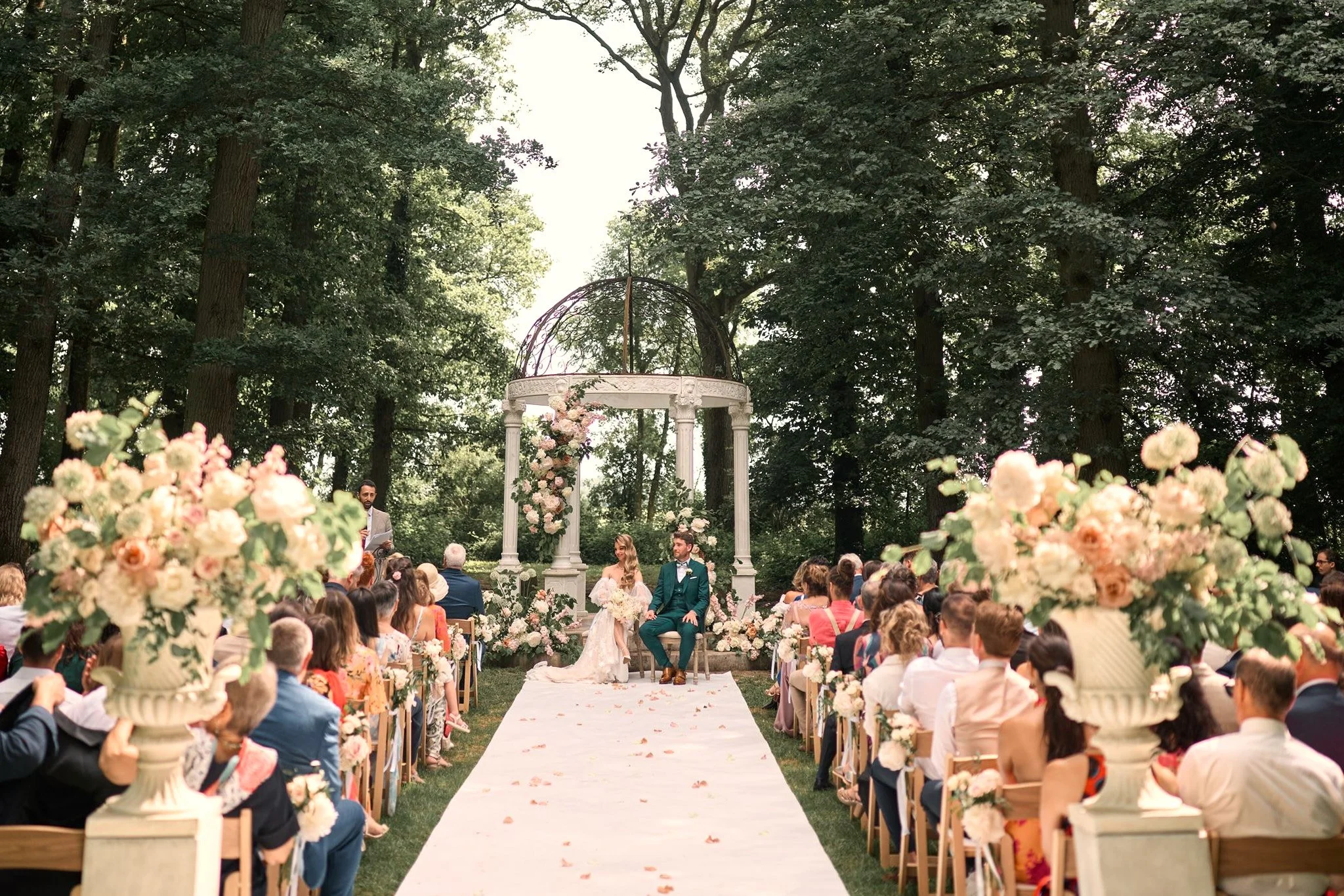 Cérémonie de mariage en plein air dans une forêt, avec un couple assis sous une arche décorée de fleurs, entouré d'invités assis sur des chaises alignées, et un officiant à gauche.