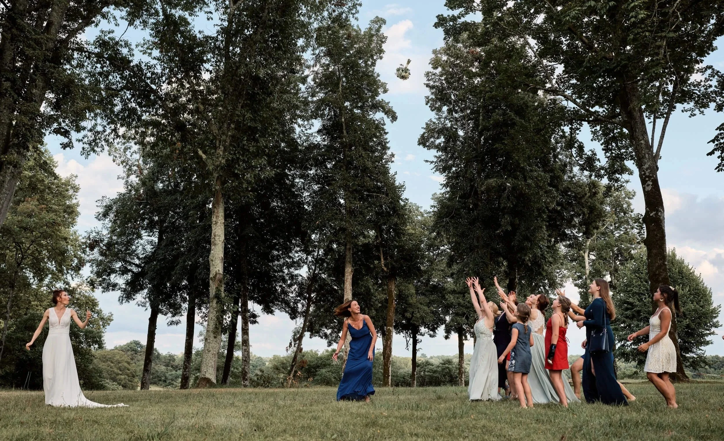 Groupe de personnes à une fête de mariage dans un champ avec des arbres en arrière-plan, la mariée en robe blanche à gauche et les invités en groupe au centre et à droite
