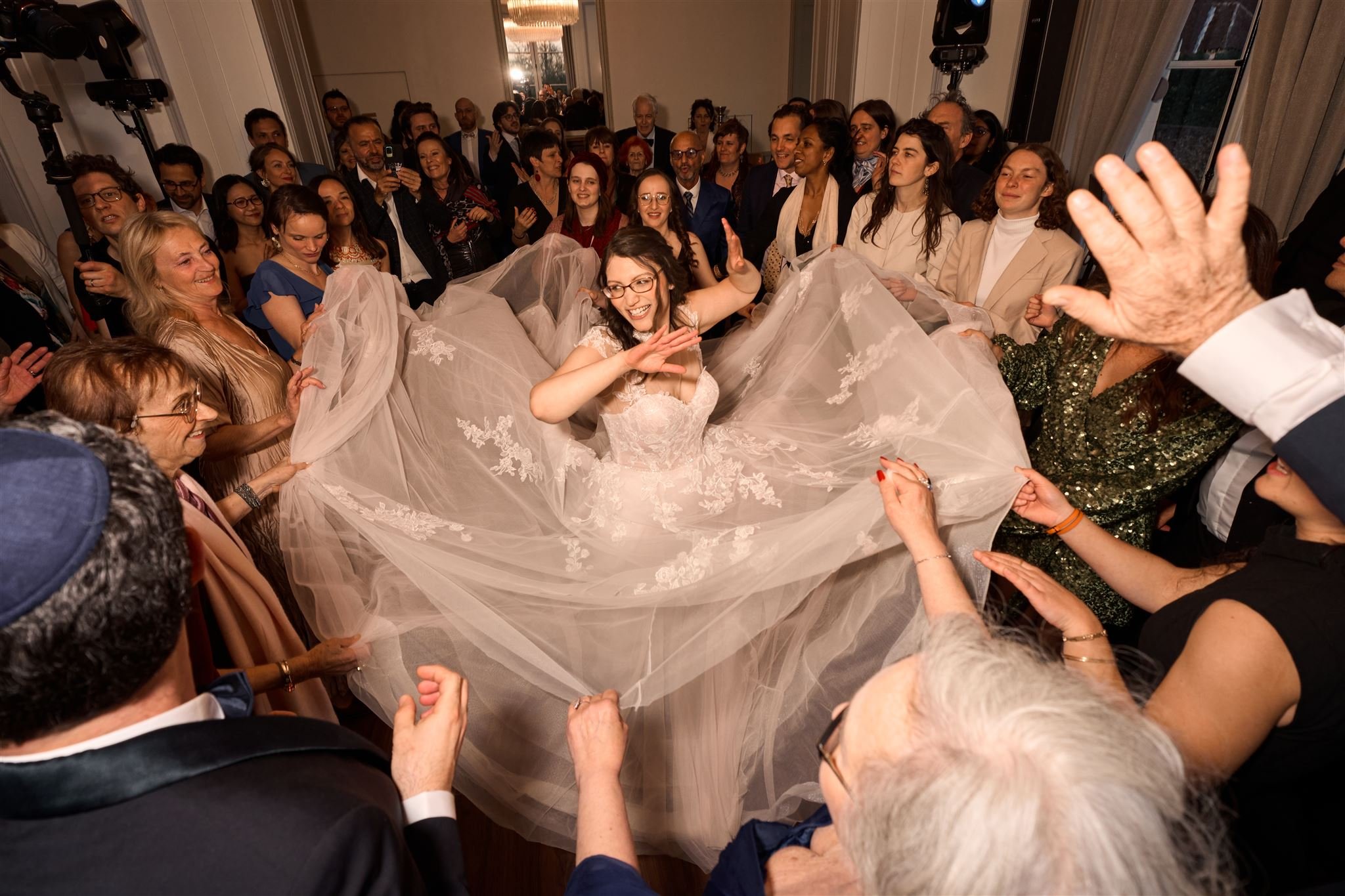 Une mariée en robe de mariage souriante gravite avec son voile en position centrale, entourée de nombreuses personnes lors d'une fête ou d'une célébration, probablement un mariage.