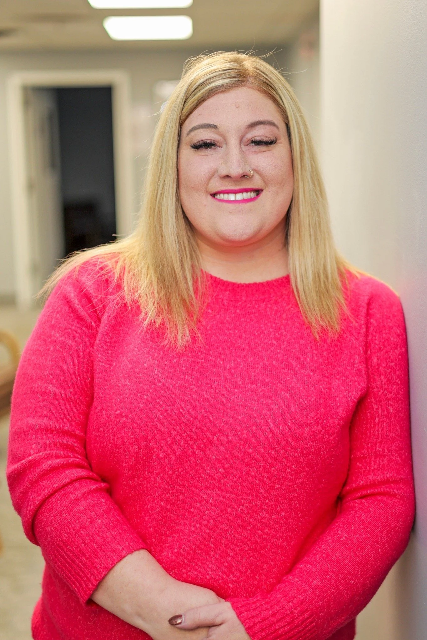 Smiling woman with curly hair, wearing a black top and necklace, in front of a purple background with silver balloons.