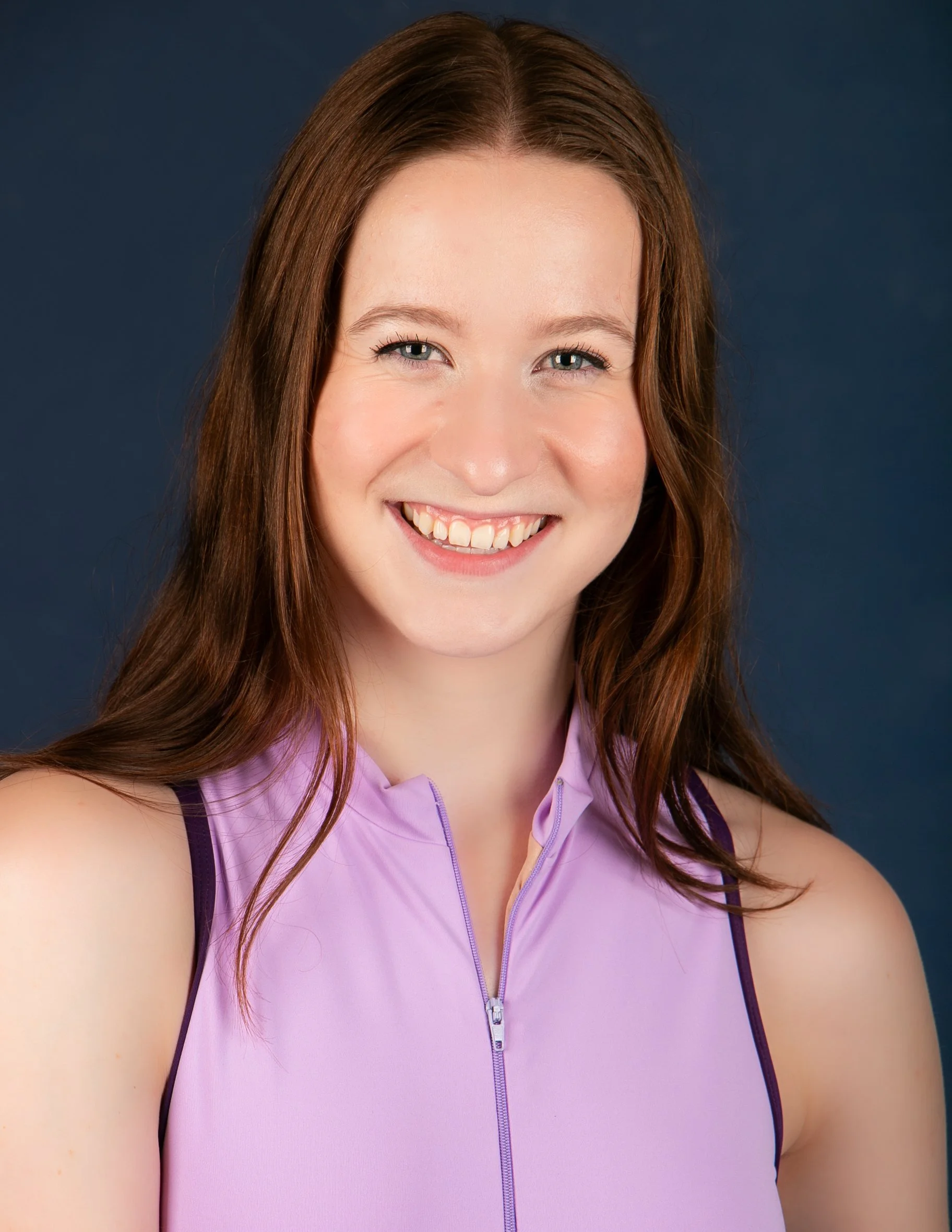 Close-up portrait of a smiling woman with long brown hair wearing a purple sleeveless athletic top with a front zipper, against a dark blue background.