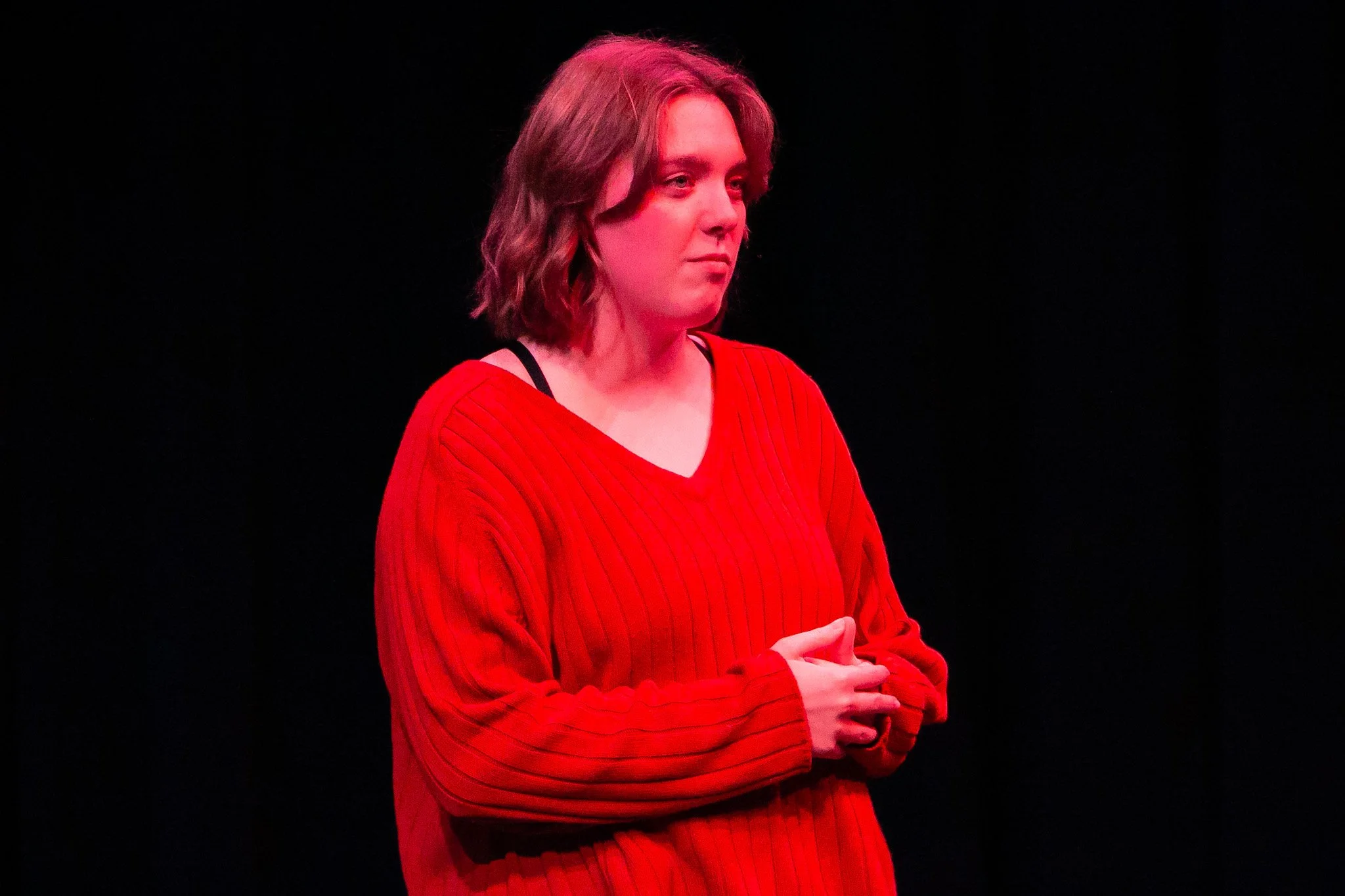 A woman with shoulder-length wavy hair wearing a red sweater with stripes, standing against a black background with her arms crossed.