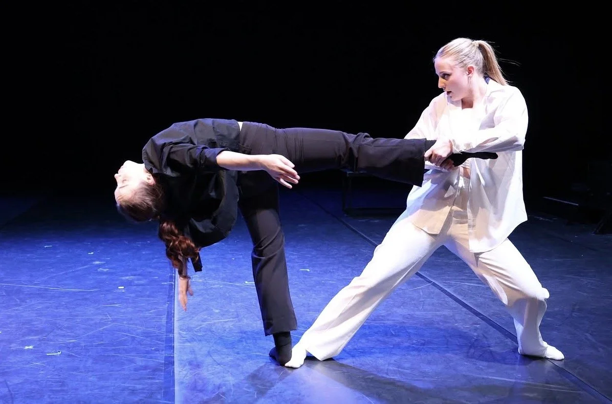 Two women are performing a theatrical or dance scene on stage, one in black and the other in white, with dramatic poses and expressions.
