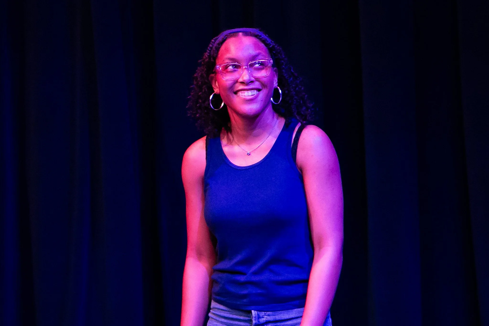 A woman smiling, wearing glasses, hoop earrings, a necklace with a small pendant, a blue sleeveless top, and gray pants, standing against a dark curtain background, illuminated by purple and pink stage lighting.