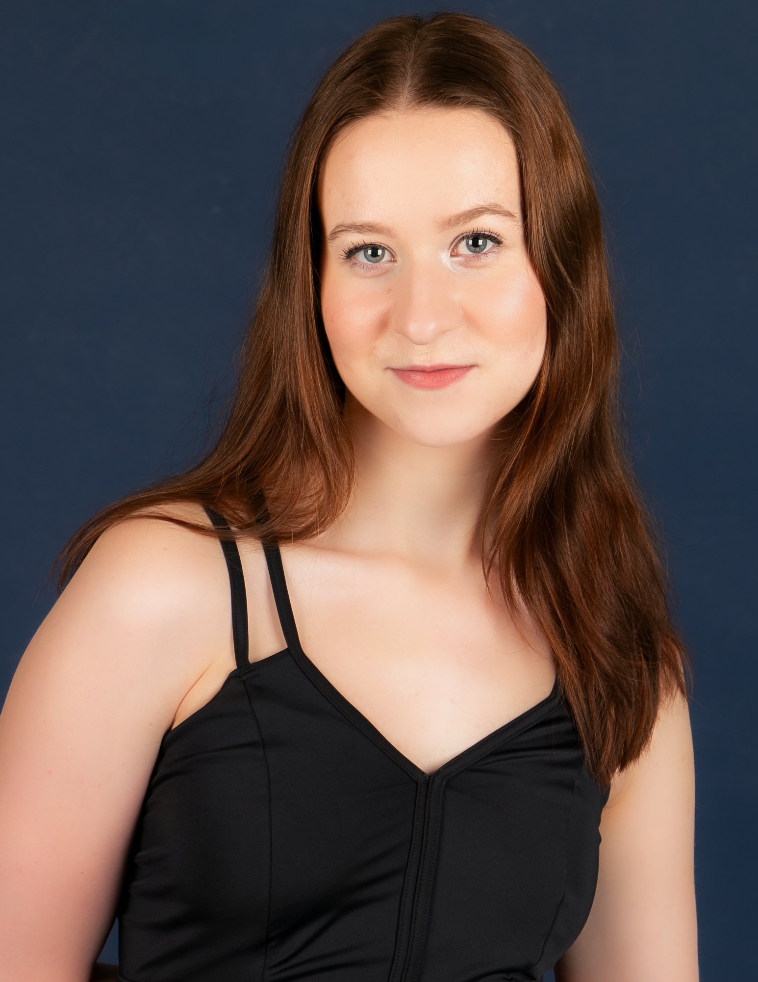 Close-up of a young woman with long, wavy auburn hair, wearing a black top with thin straps, smiling softly with a dark blue background.