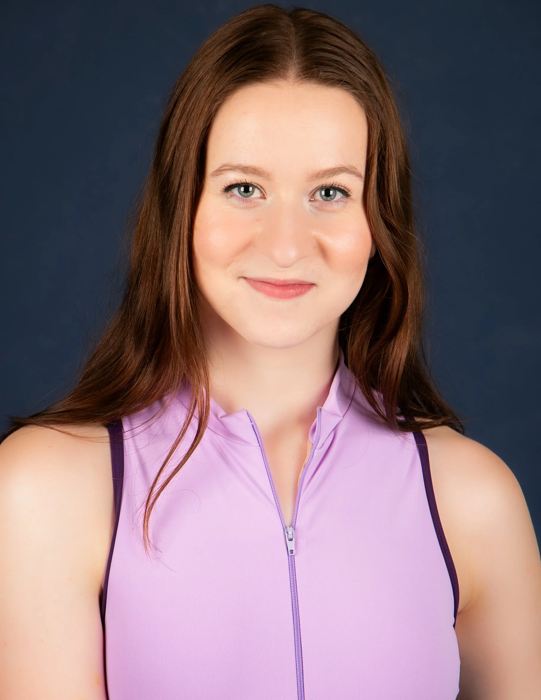 A woman with long, wavy red hair wearing a sleeveless purple athletic top with a zipper in front, set against a dark background.
