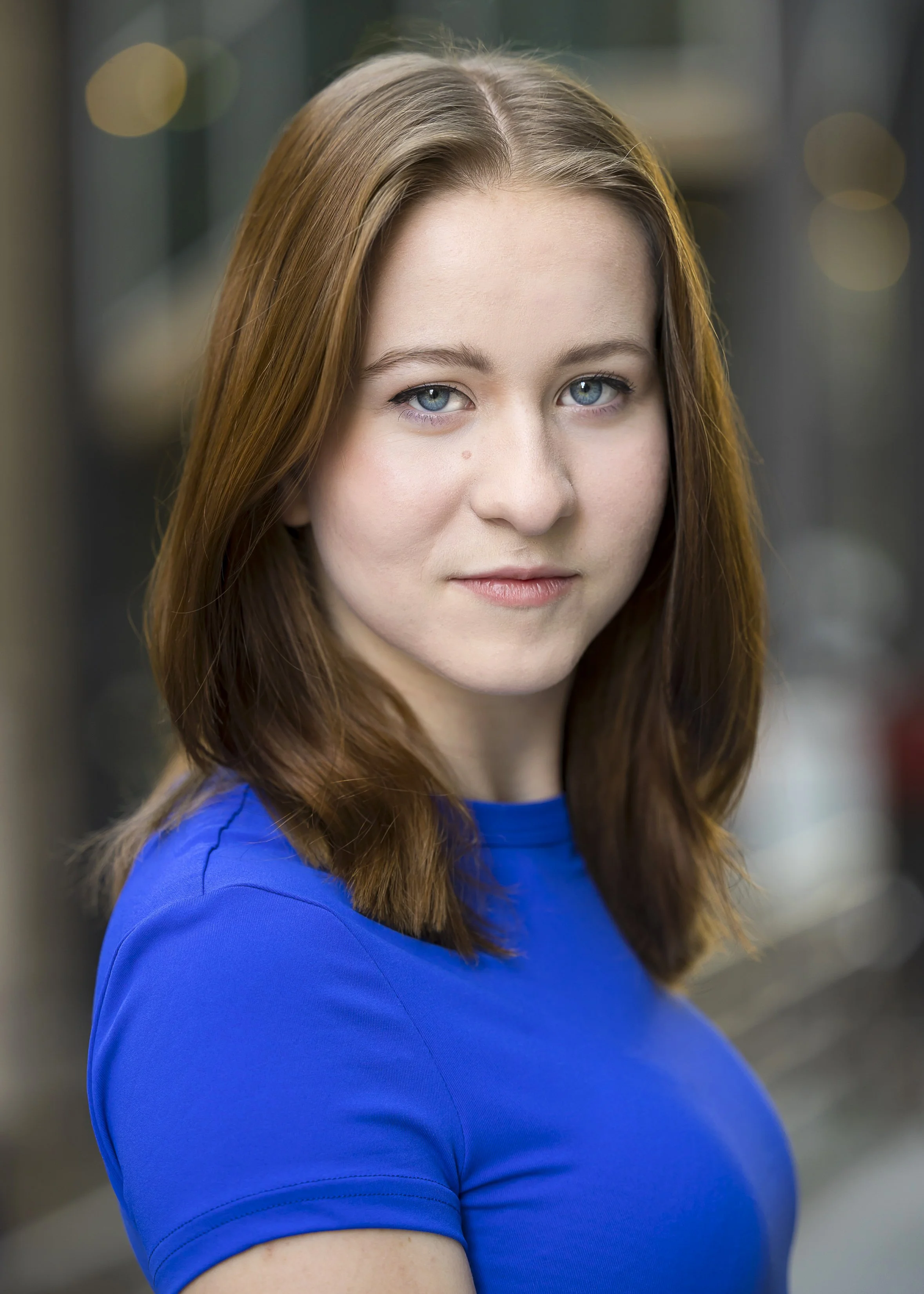 Close-up of a woman with long, light brown hair, blue eyes, wearing a royal blue top, standing outdoors with a blurred background.