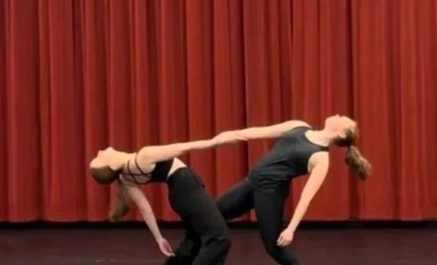 Two women performing an acro yoga pose on stage with red curtains in the background, one bending backwards and the other leaning forwards to support her.