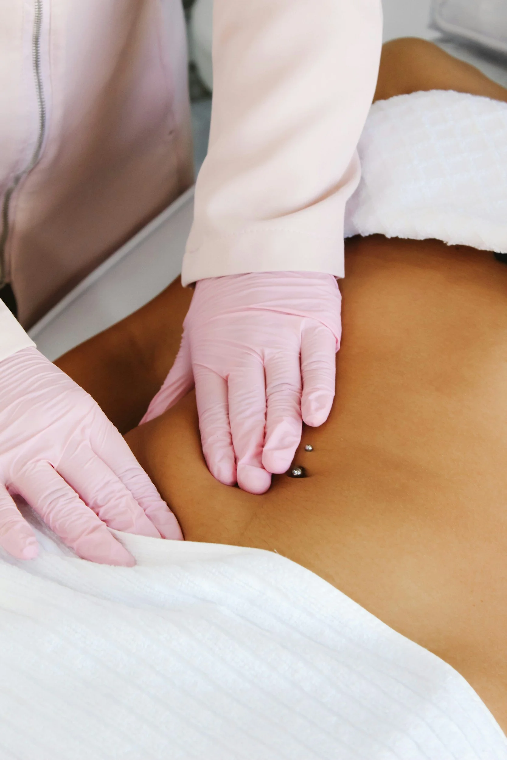 A healthcare professional wearing pink gloves is inserting a needle into a patient's belly button area, with a white towel covering part of the patient’s abdomen.