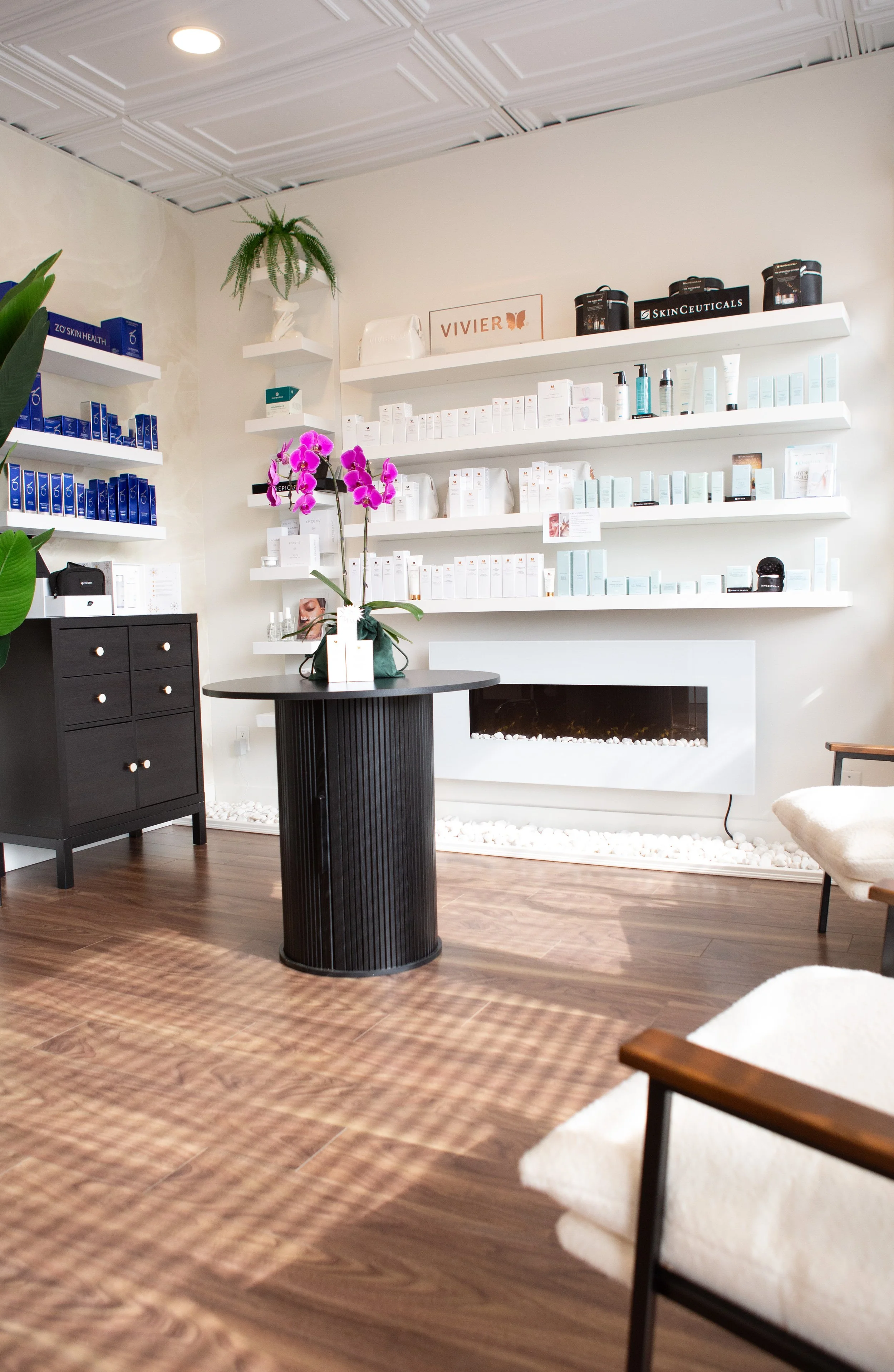 Interior of a modern beauty or skincare spa with white shelves holding skincare products, a table with a potted orchid, chairs, and a wall-mounted fireplace.