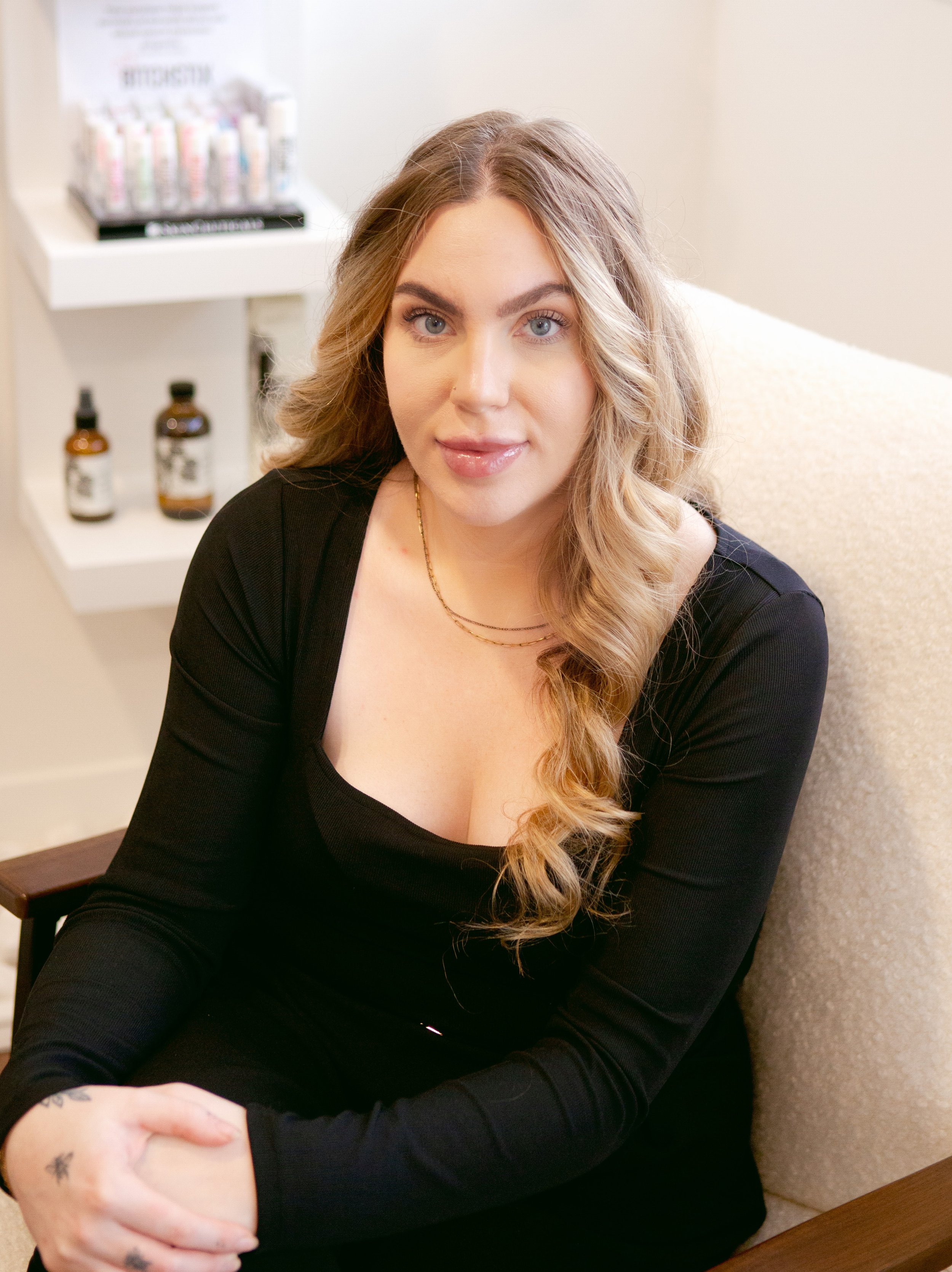 A woman with long, wavy blonde hair, wearing a black top and gold necklaces, sitting on a beige armchair in a room with a white shelf holding small bottles and a stack of books in the background.