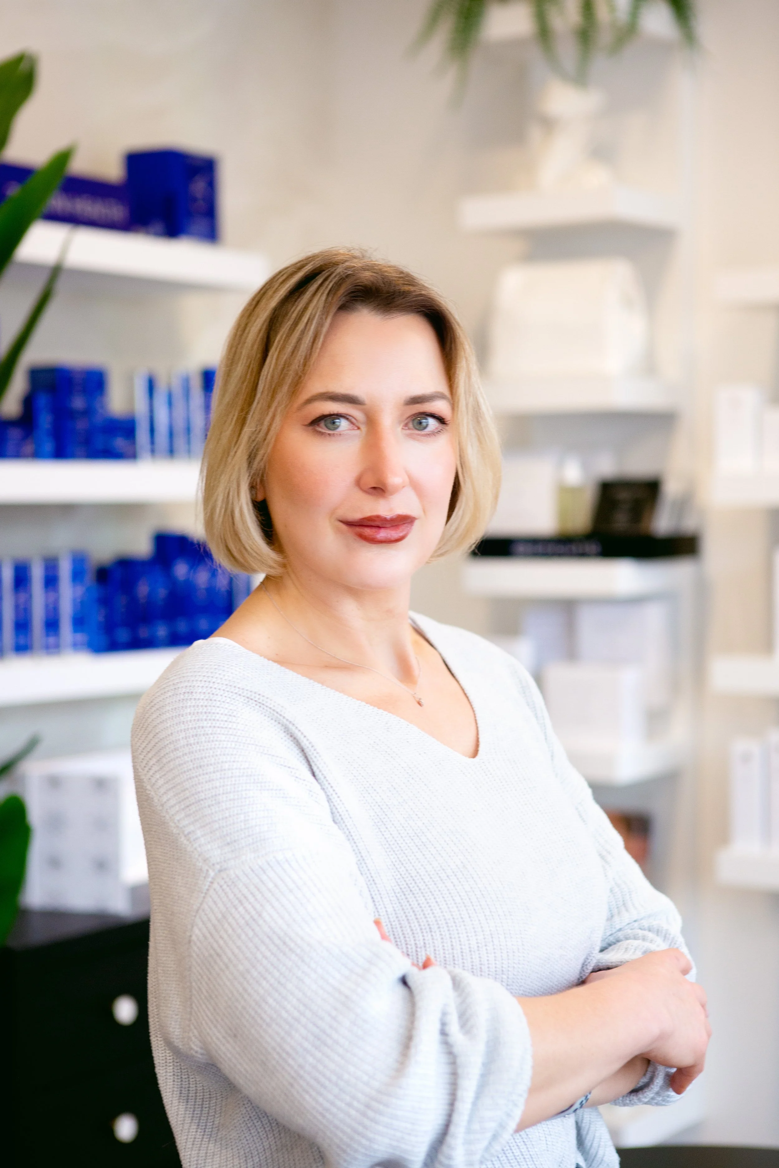 A woman with shoulder-length blonde hair and blue eyes, wearing a light gray sweater, standing in an indoor setting with white shelves and blue boxes in the background.