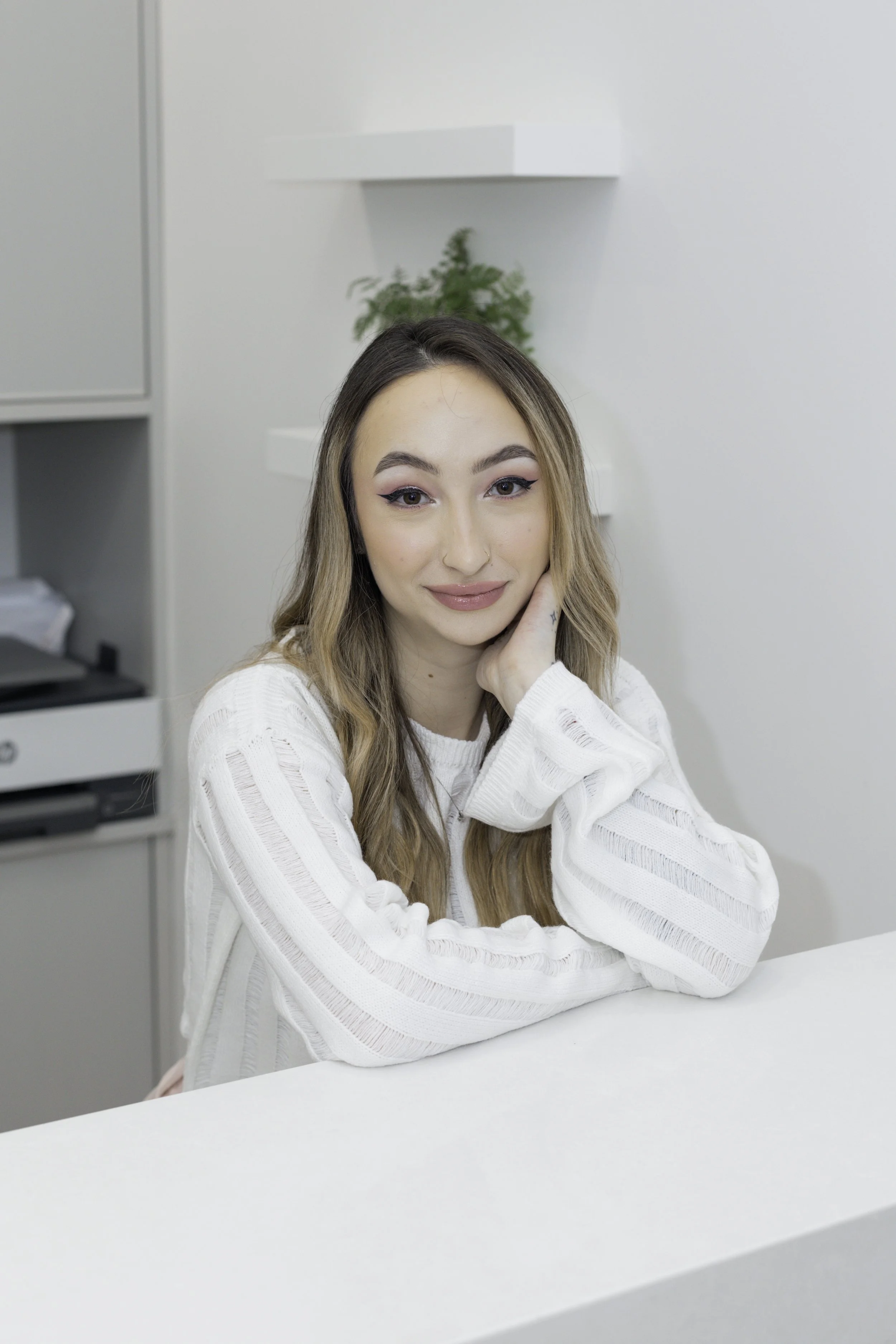 Young woman with long wavy hair, wearing a white sweater, sitting at a white counter in a bright, minimalist room with a plant and shelf in the background.