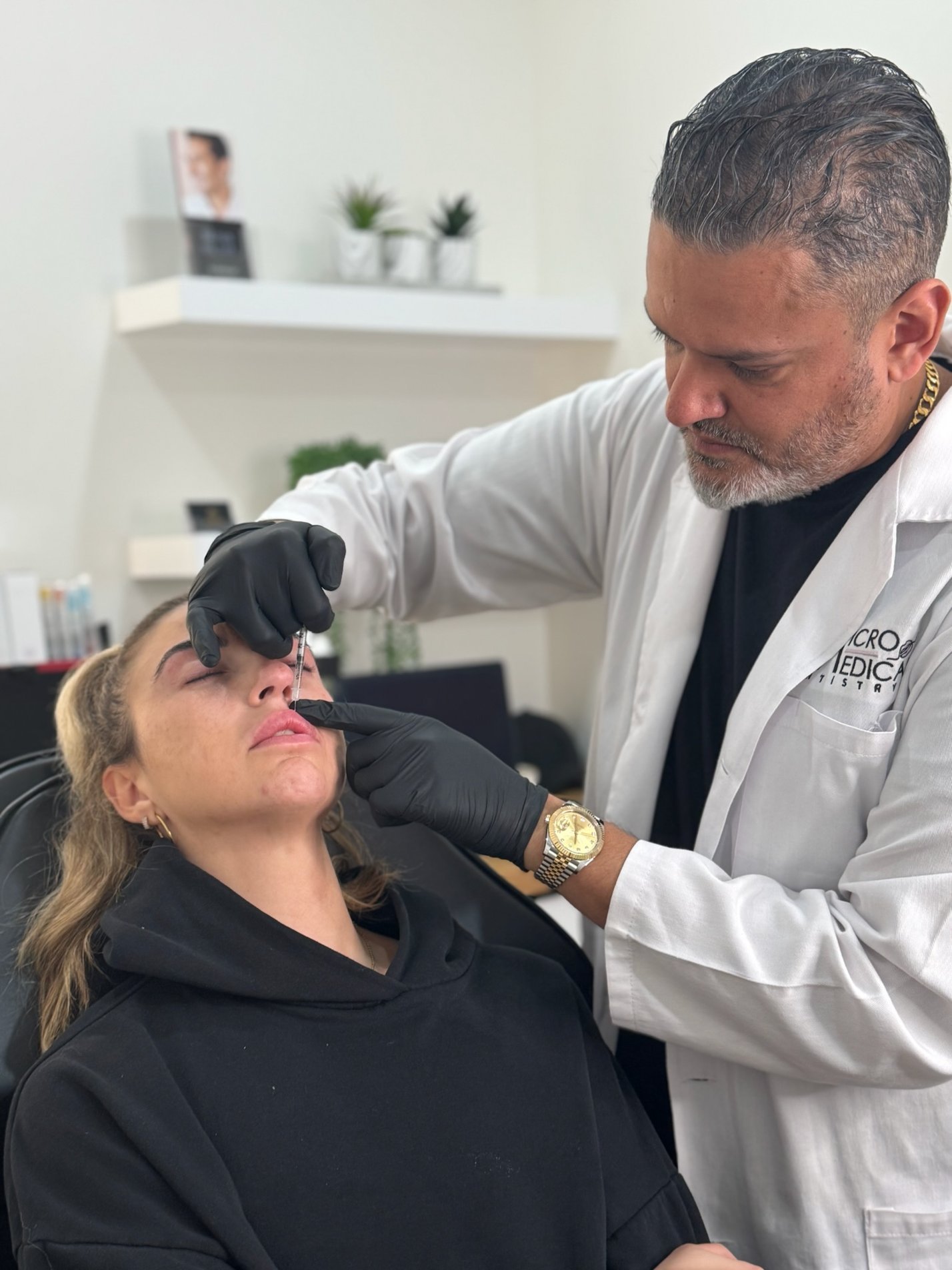 A medical professional with dark hair and a gray beard, wearing a white lab coat and black gloves, administering botox to a woman with blonde hair, who is reclined in a chair with her eyes closed in a clinical setting.