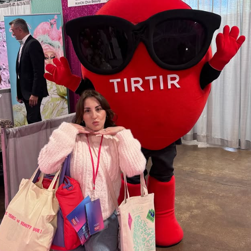 A young woman posing with a large anthropomorphic heart mascot wearing sunglasses and red gloves at an indoor event.