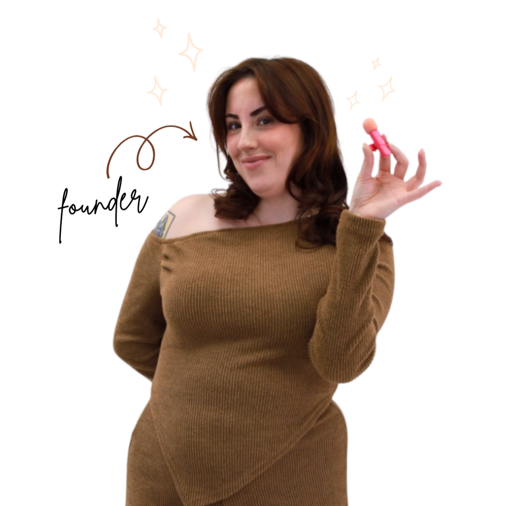 A woman with brown hair, wearing a brown off-the-shoulder sweater, holding a pink pill bottle in her right hand, smiling at the camera.