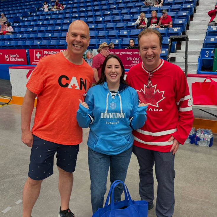 Three people standing together in an arena with blue seats in the background. The person on the left is a man in an orange Canada T-shirt and shorts, smiling. The person in the middle is a woman in a blue Downtown Sudbury hoodie, smiling and making a gesture with her hands. The person on the right is a man in a red Canada hockey jersey, smiling. There are spectators seated behind them and water bottles on the ground.