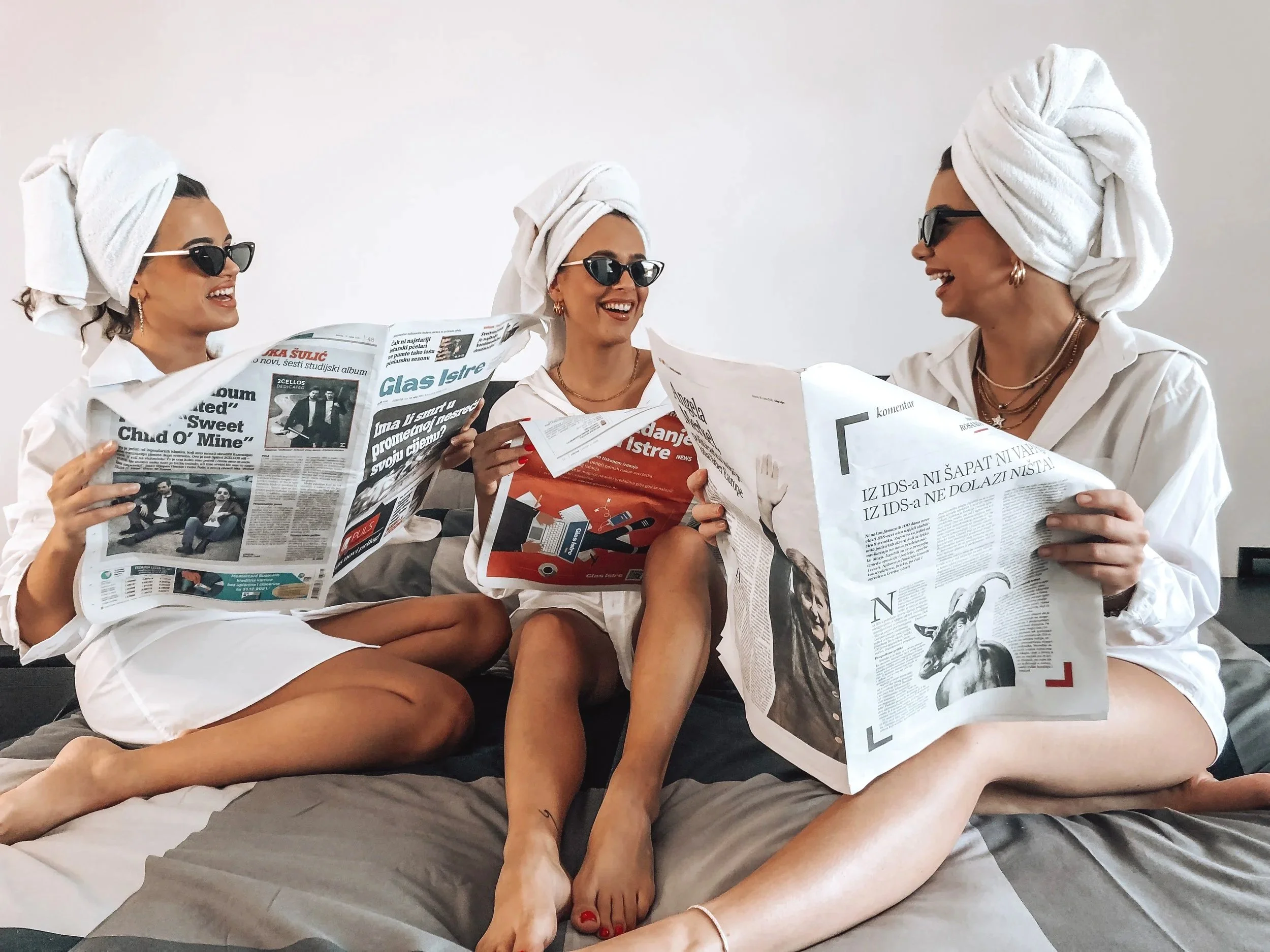 Three women with hair wrapped in white towels, wearing sunglasses and gold jewelry, are sitting close together and smiling while reading newspapers.