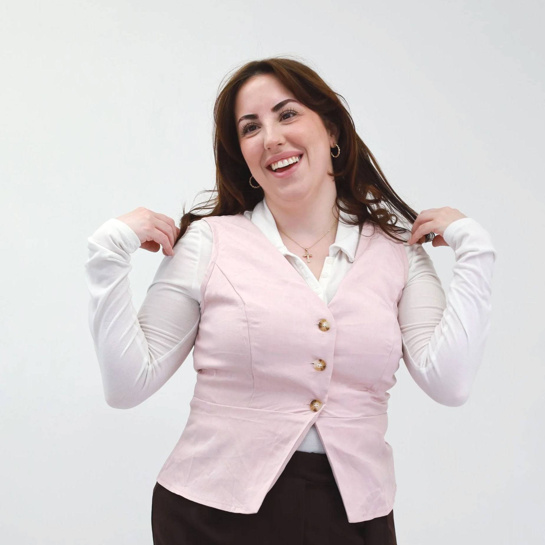 A woman with brown hair smiling, wearing a white blouse and a pink vest, adjusting her hair with both hands, standing against a plain white background.