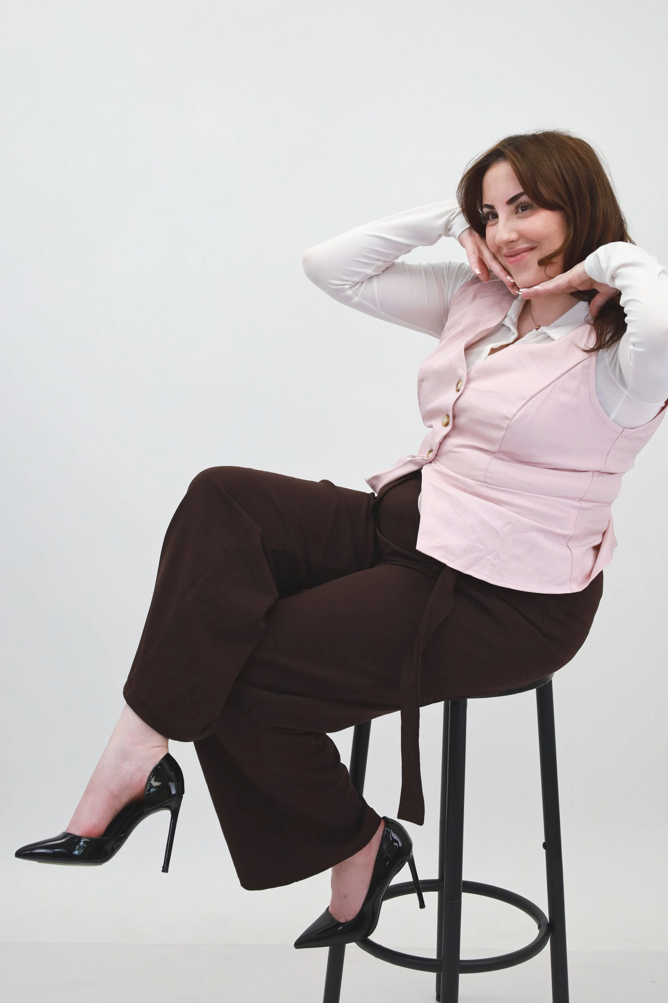 Woman sitting on a high stool, wearing a light pink vest over a white shirt, dark brown pants, and black high heels, posing with her hands framing her face against a plain white background.