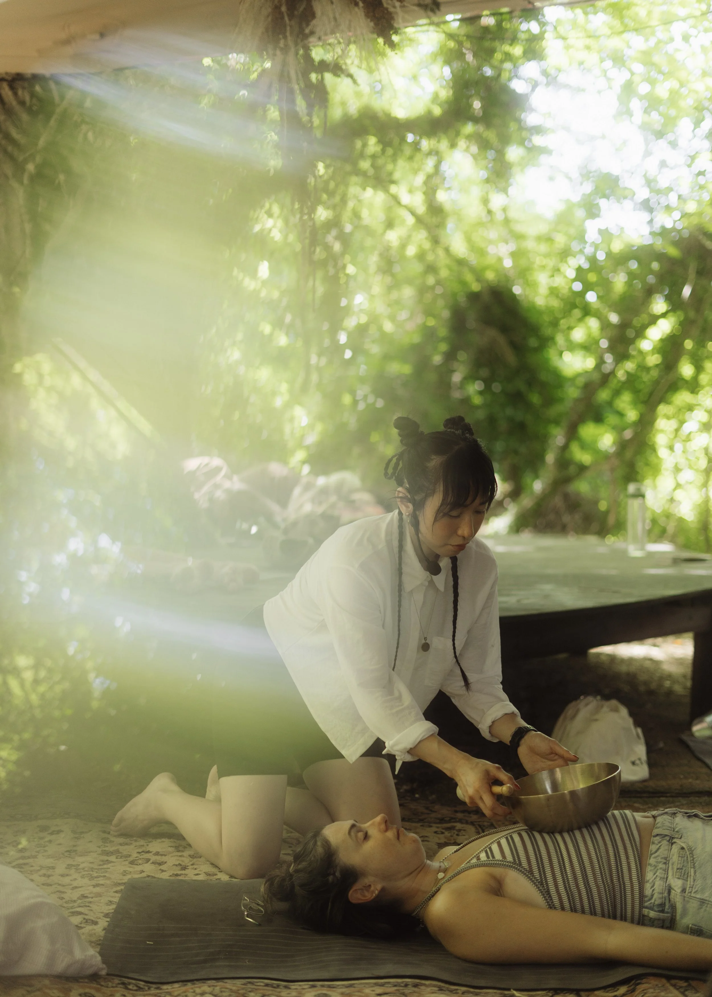 Sound mirror plays a singing bowl on the body of a young woman who is lying on a yoga mat with her eyes closed. They are outdoors at a festival, in the background are trees and dappled sunlight.