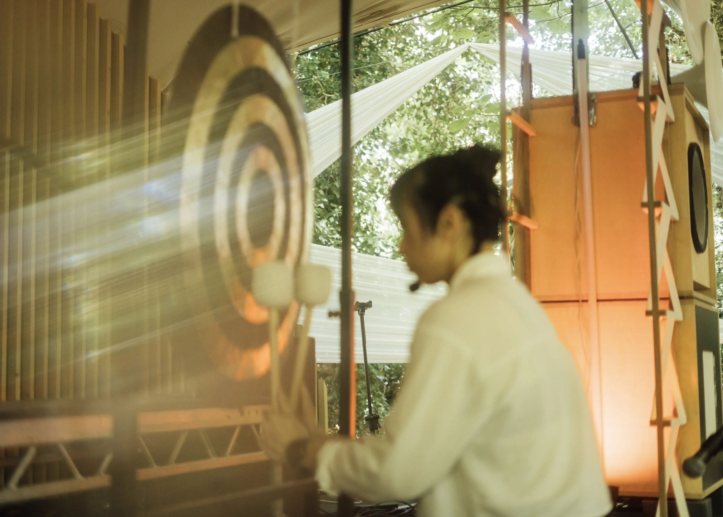 Sound Mirror is playing a gong, holding two wool mallets in front of large speakers. The image is blurred by light and motion.