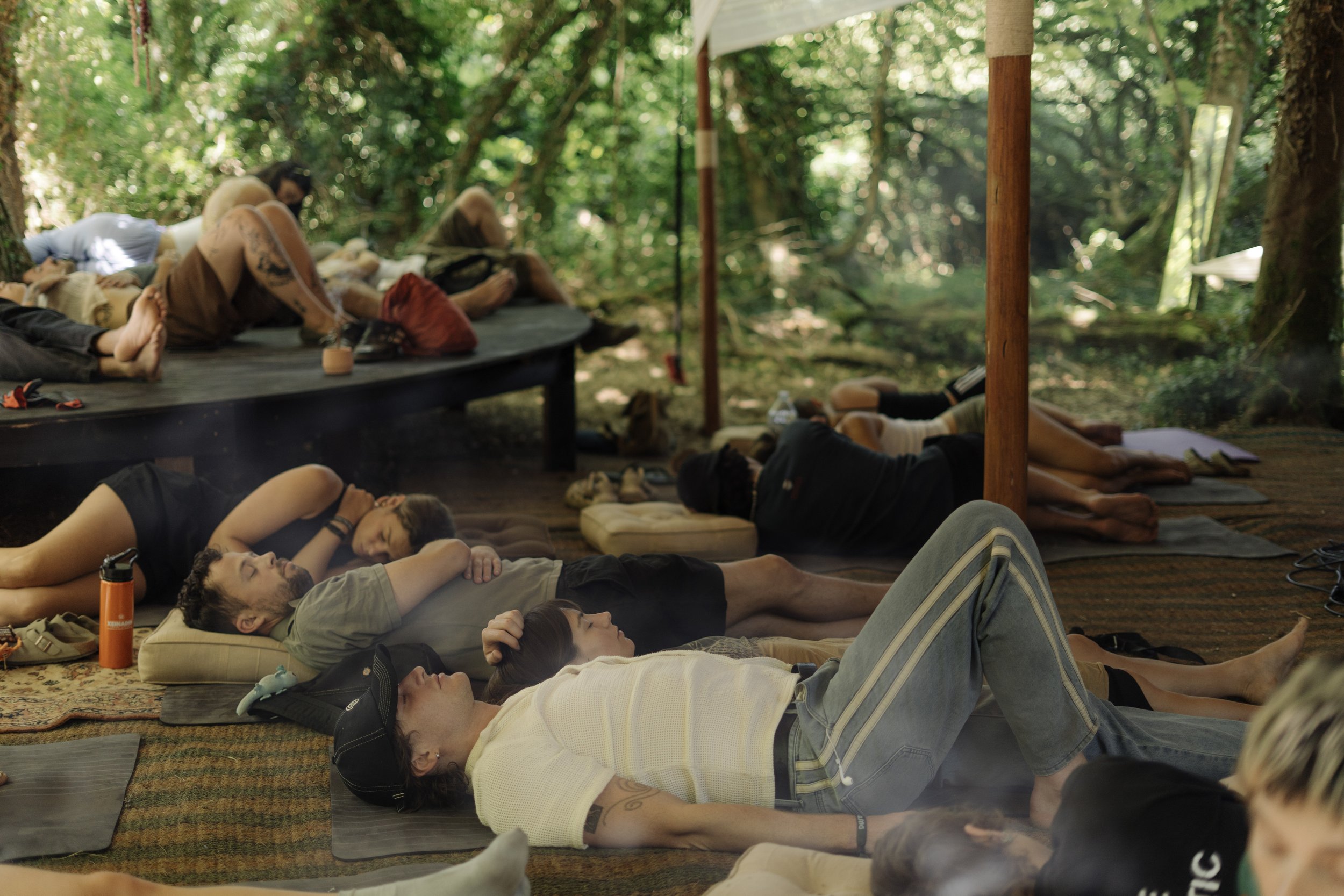 People are laying down with their eyes close on yoga mats looking relaxed. They are outdoors at a festival. The area is surrounded by trees and dappled sunlight.
