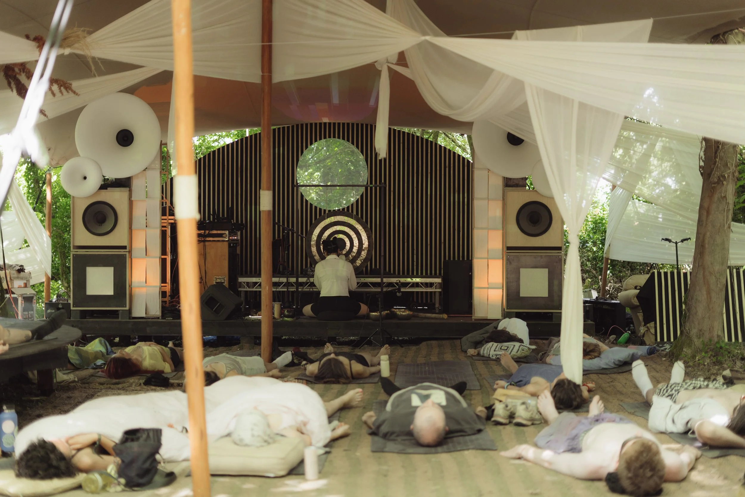 Sound Mirror playing a gong on a stage flanked by tall speakers with space-age looking cones in front of a group of people lying down listening. It is an outdoor stage at a festival with a fabric draped canopy surrounded by trees and sunlight.