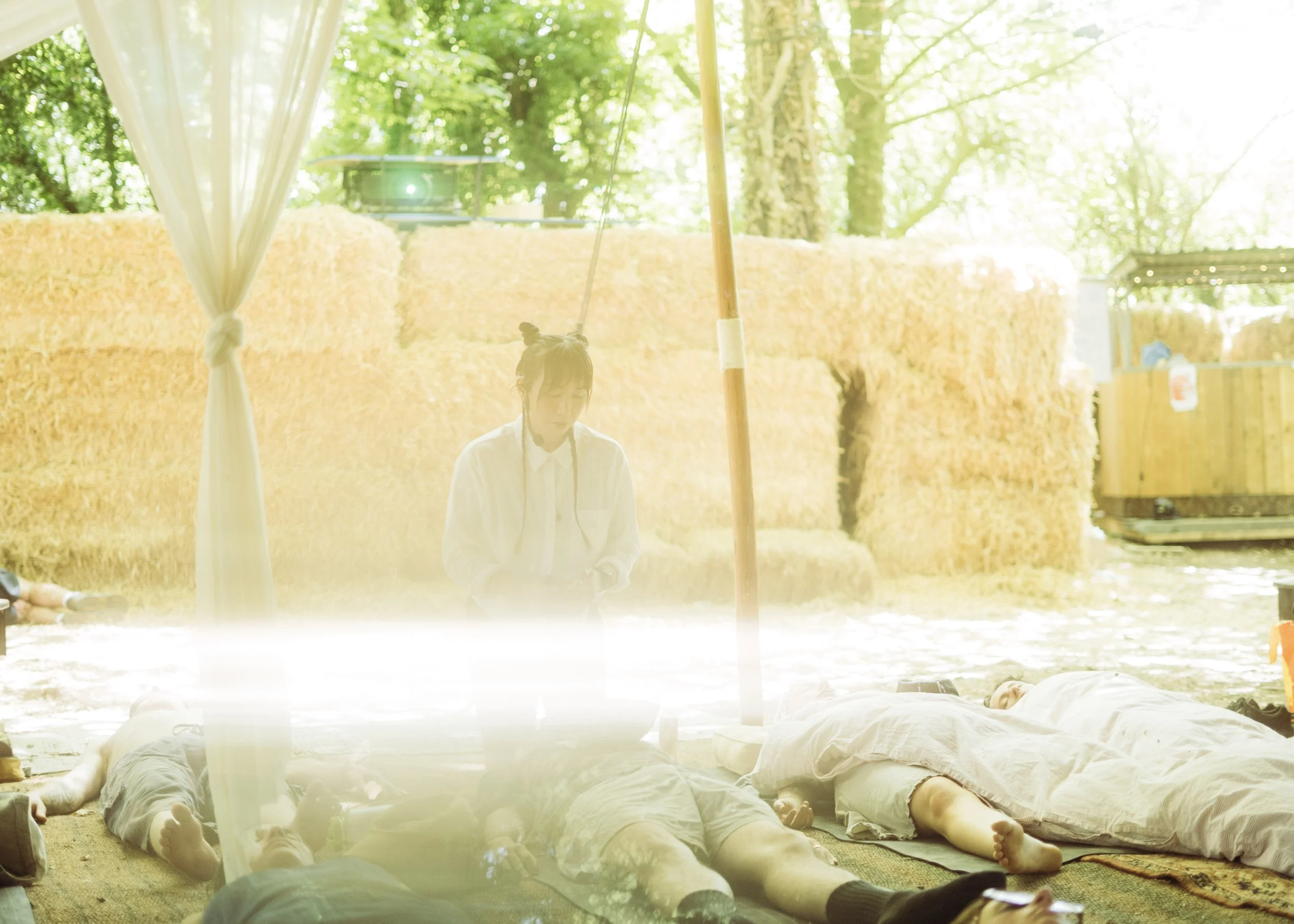 Sound Mirror is playing sound bath instruments on people laying down on yoga mats looking relaxed. The photograph is sun bleached and obscured by a flash of bright light. They are outdoors at a music festival. In the background are straw bales.