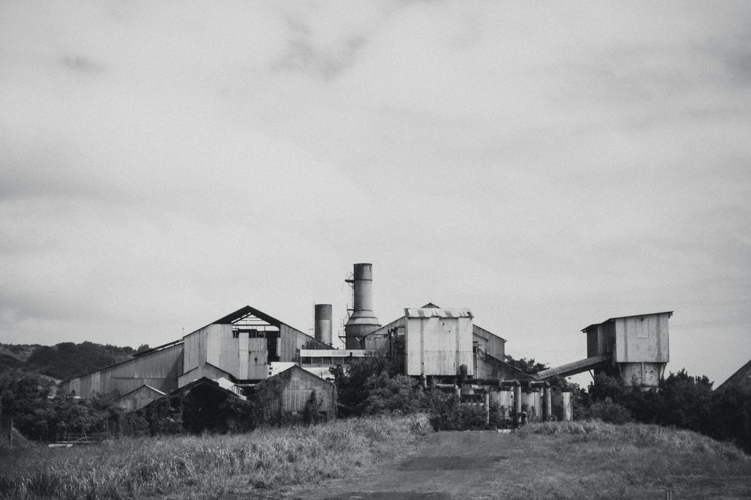 Black and white photo of an abandoned factory with dilapidated buildings and smoke stacks in a rural area.
